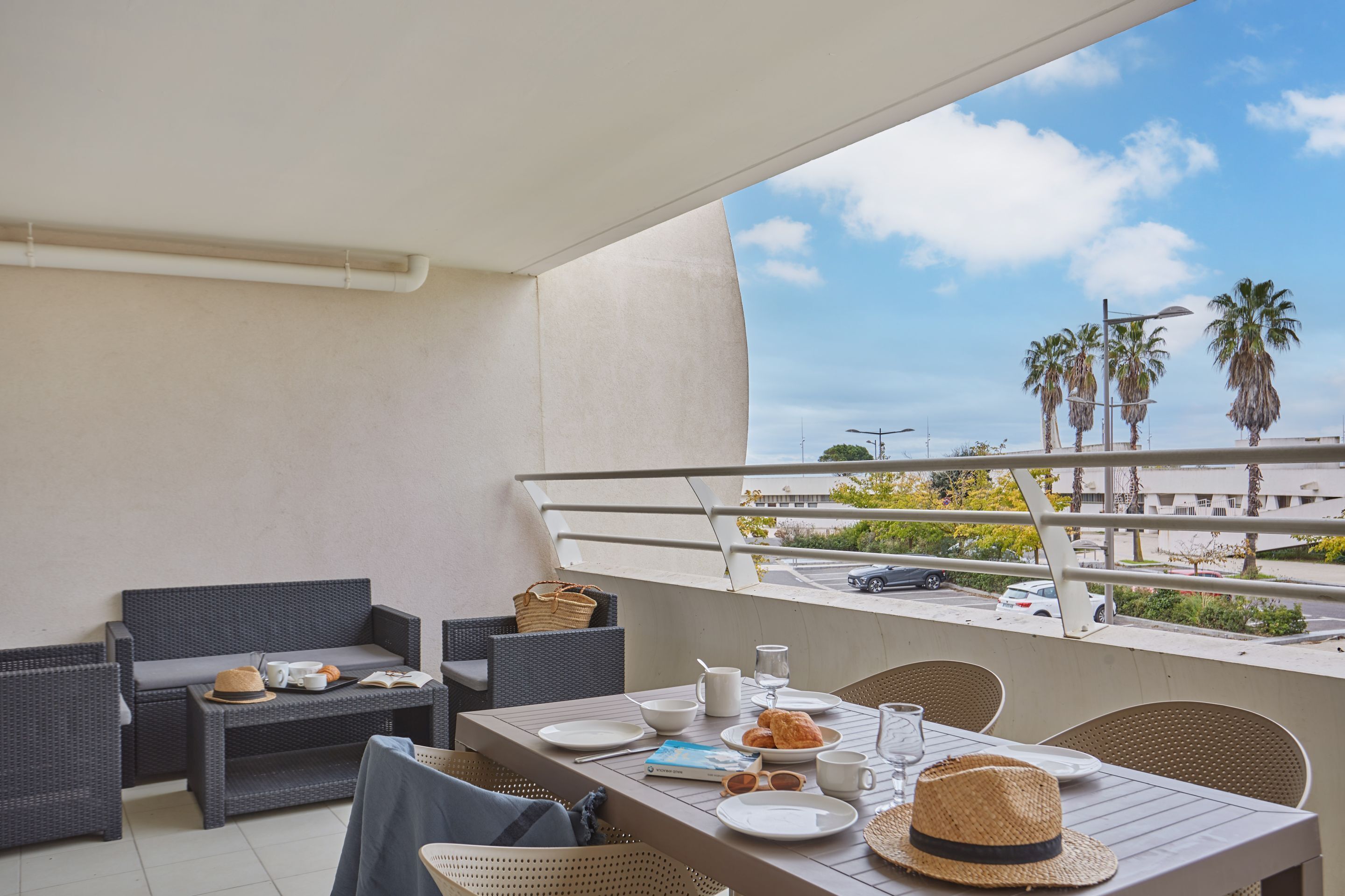 Varanda com mesa posta para o café da manhã, móveis de vime e vista para palmeiras e uma rua.
