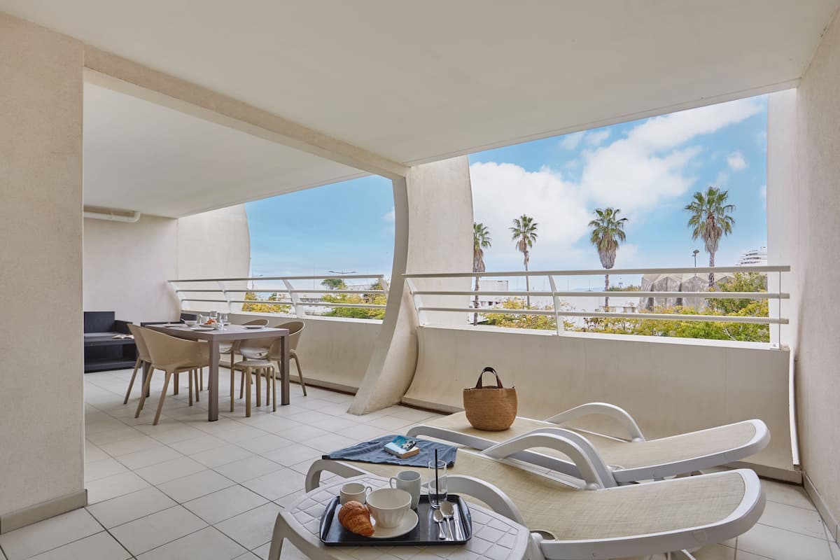 Een balkon met een eettafel, stoelen en een ontbijttafel. Achtergrond met palmbomen en een heldere lucht.