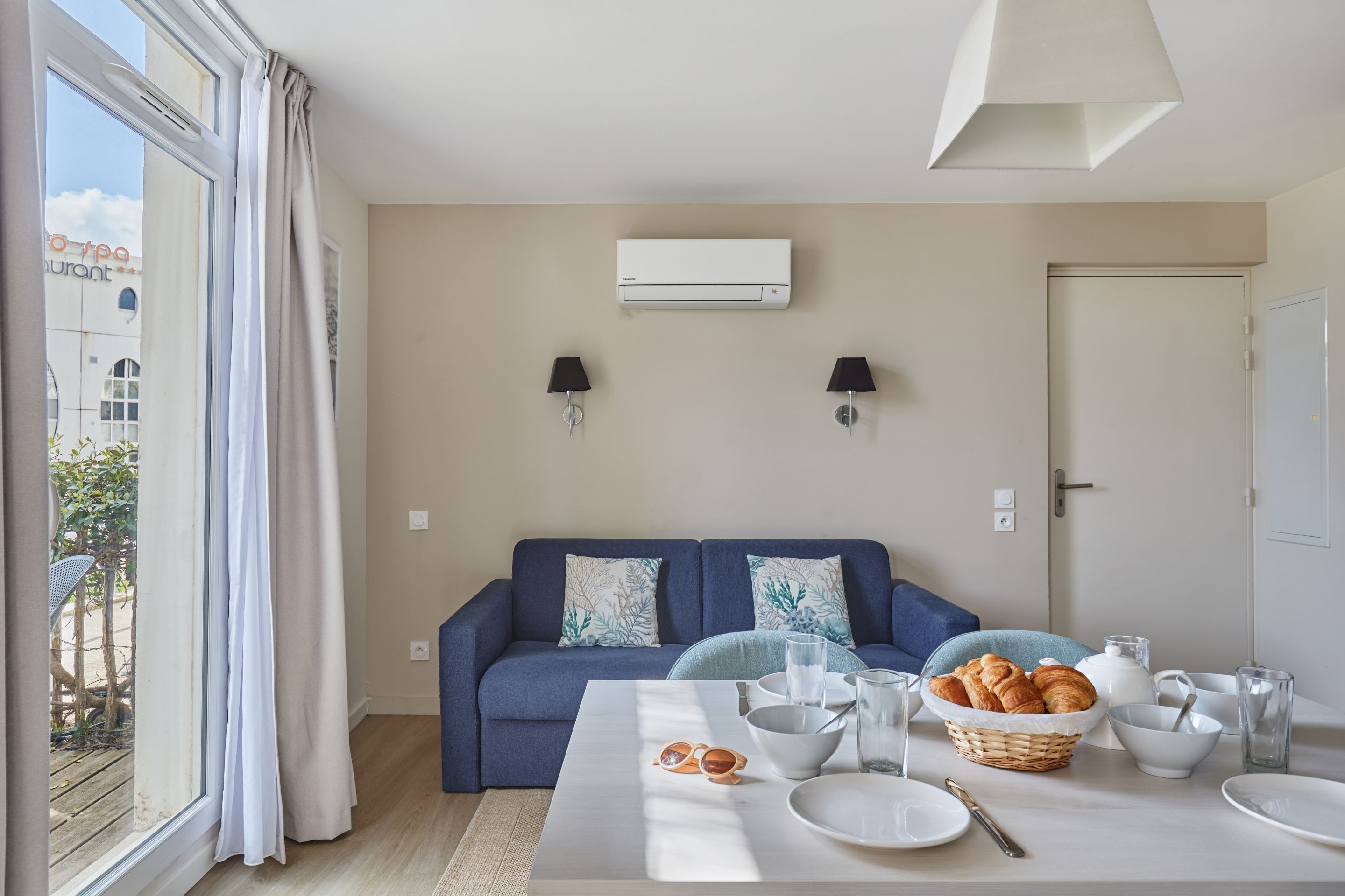 A modern living room with a blue sofa, a dining table with breakfast items, and a large window with curtains.