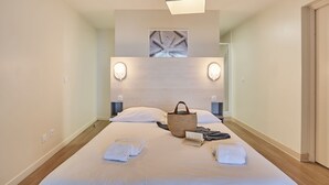 Un lit bien fait, avec des serviettes, un livre et un sac, dans une chambre d’hôtel moderne dotée d’une tête de lit en bois et de lampes fixées au mur.