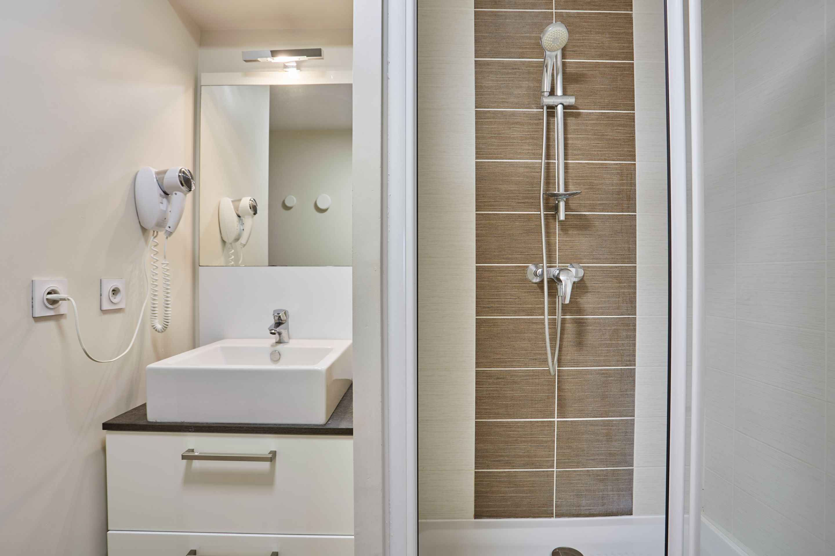 Casa de banho com lavatório branco, espelho, secador de cabelo e chuveiro com parede revestida a azulejos castanhos.