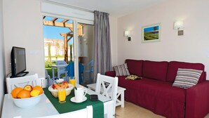 A living area with a red sofa, a white dining table with a bowl of fruit and a glass of orange juice, a TV, and a view of a garden through a sliding door. - Résidence Odalys Le Mas des Alpilles (Paradou)