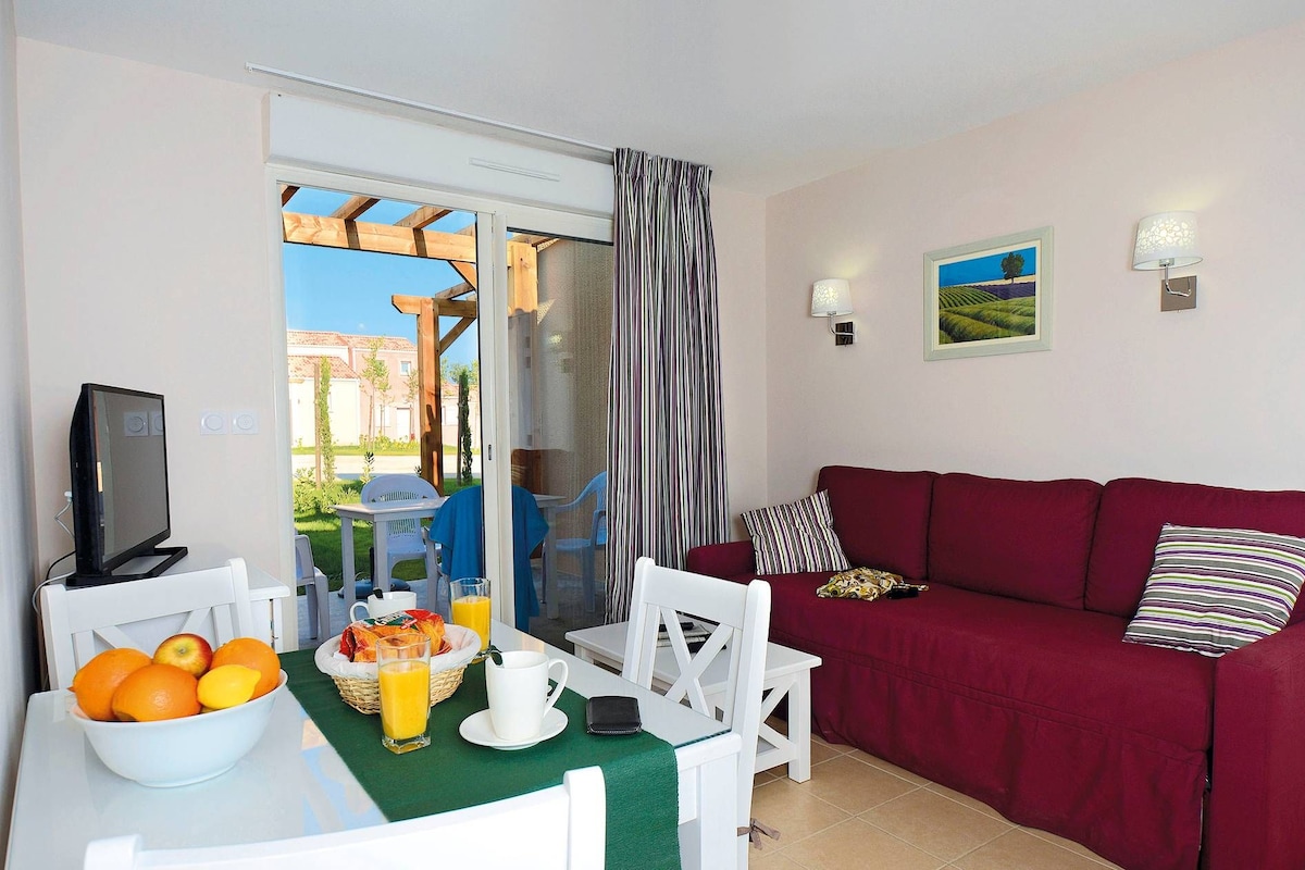Una sala acogedora con un sofá rojo, una mesa de comedor con fruta y jugo, un televisor y vistas a través de un ventanal.