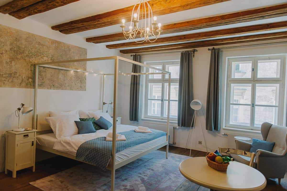 A bedroom with a four-poster bed, a bedside table with a lamp, a chair, a round table with a fruit basket, and a view of the outside through a window.
