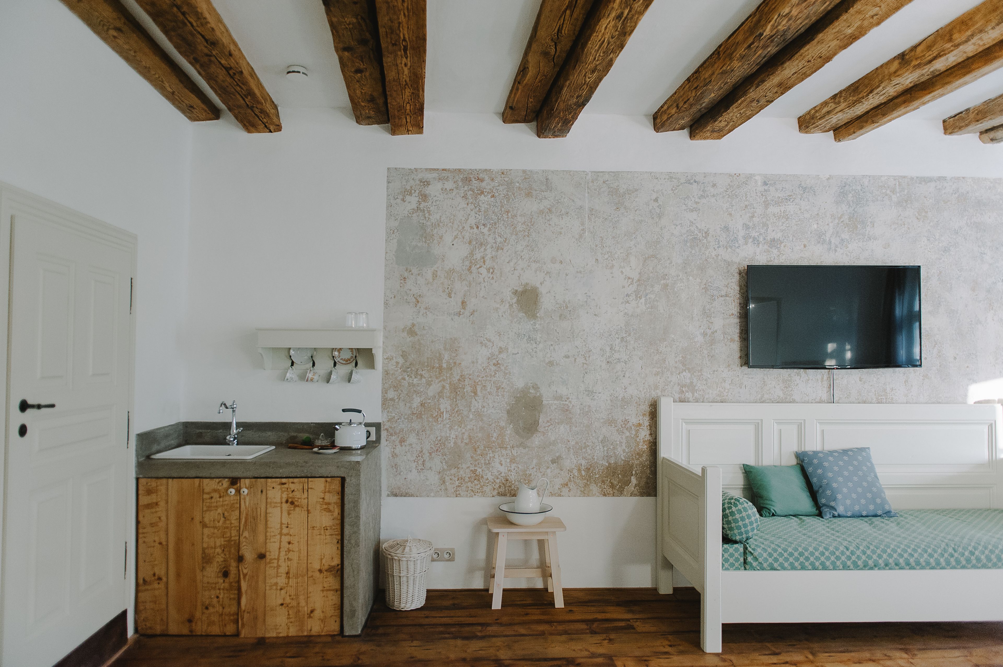 Une pièce avec un plafond en bois, un lit recouvert d’une couverture verte, une commode blanche, un petit tabouret et un téléviseur fixé au mur.