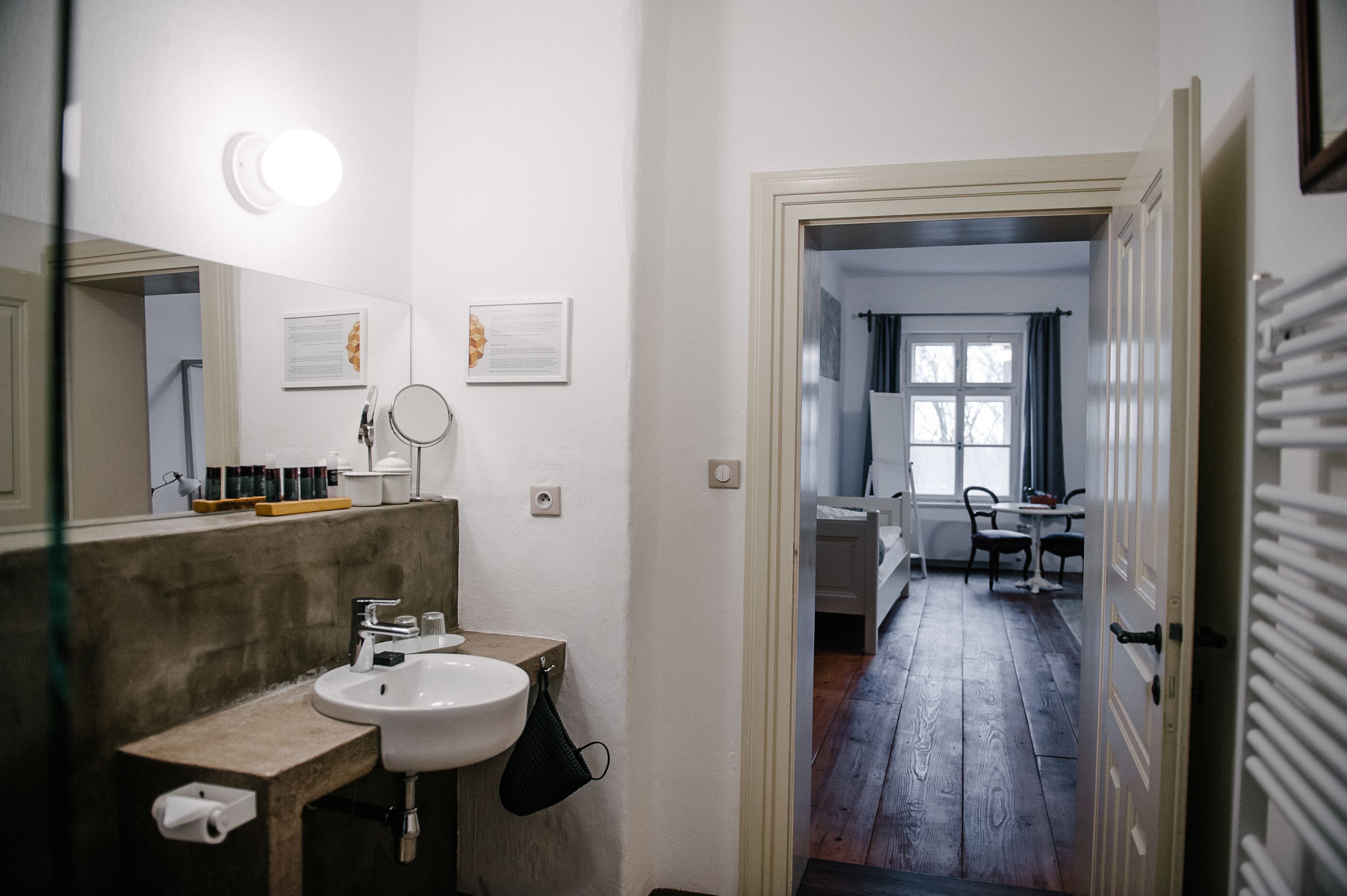 A bathroom with a sink, mirror, and toiletries. There is a hallway leading to a bedroom with a window and a view of the outside.