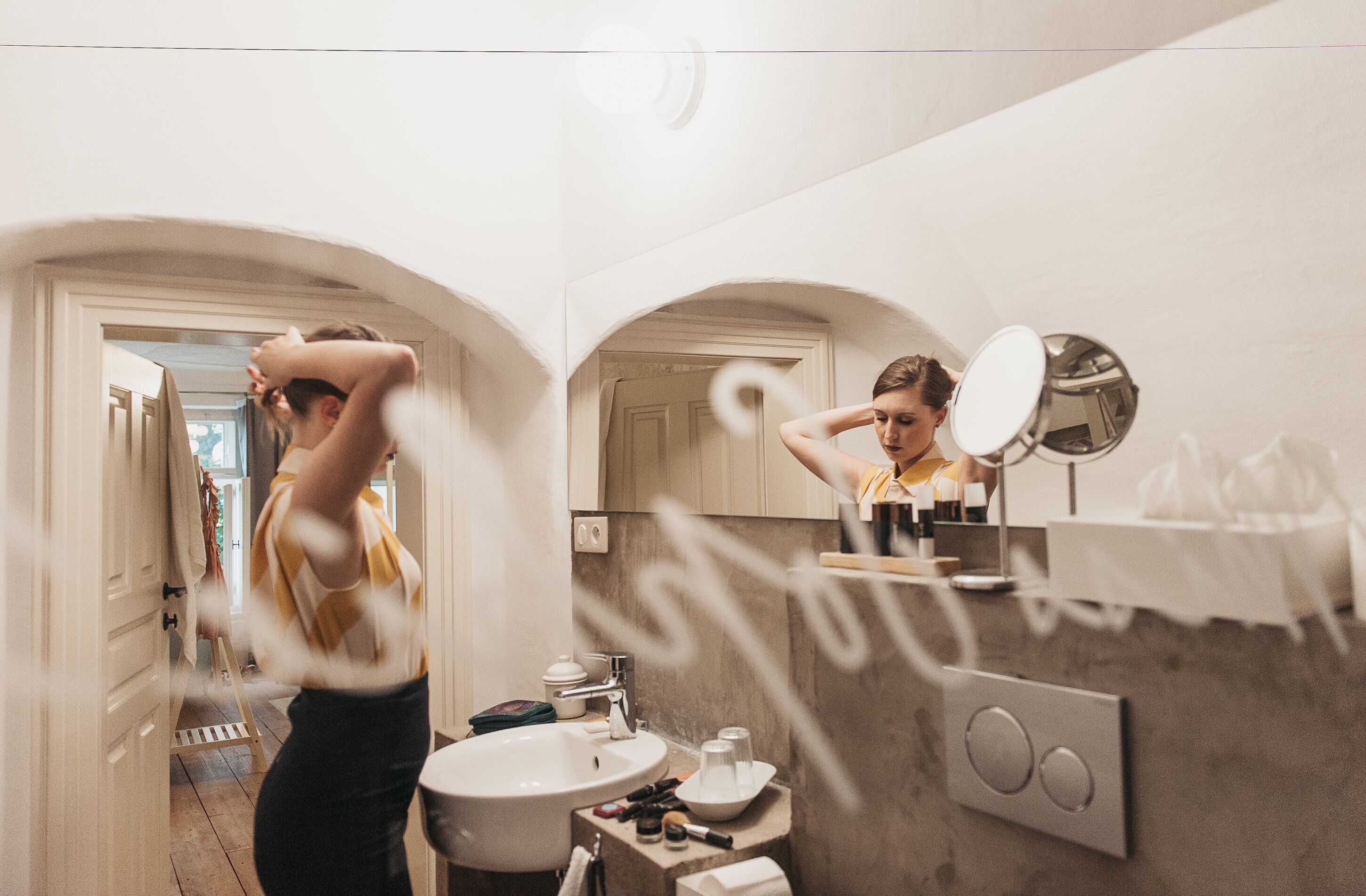 Un baño con lavamanos, espejo y tocador. Hay una puerta que da a otra habitación.