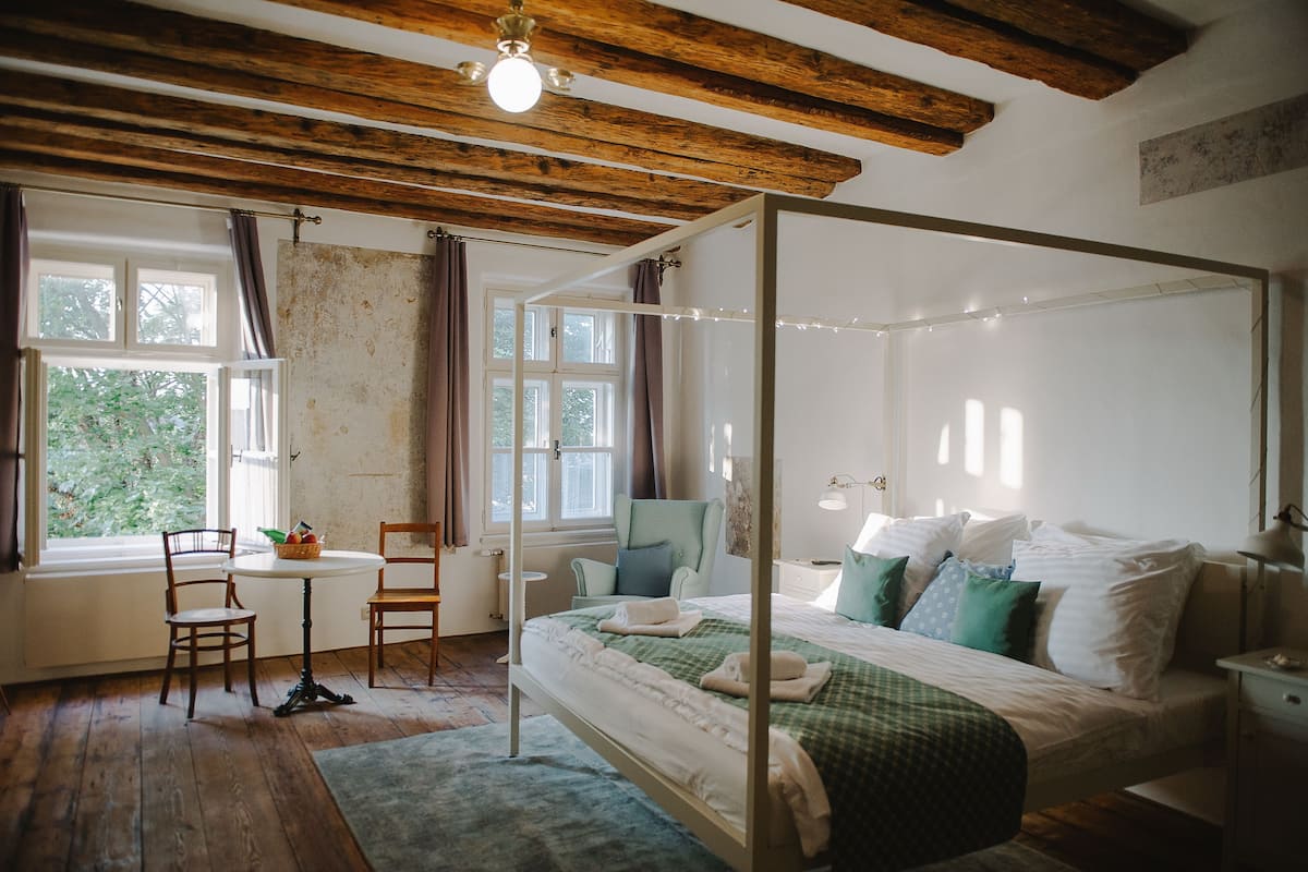 A four-poster bed with white linens, a green checkered bedspread, and multiple pillows. There is a small round table with chairs and a basket of fruit. The room has wooden floors and a window with curtains.