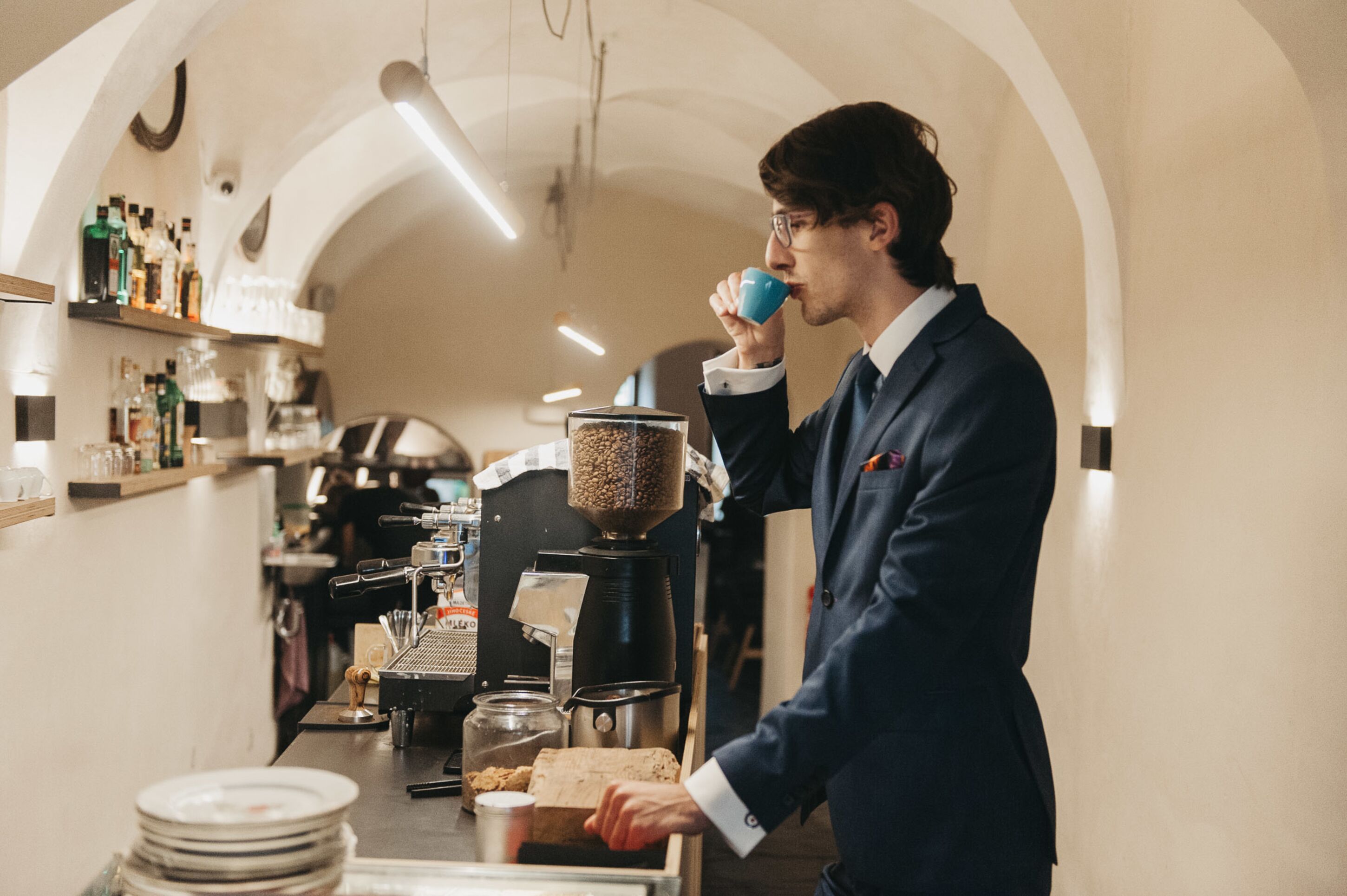 A barista in a dark suit preparing coffee in a well-lit, arched room with shelves of bottles and a coffee machine.