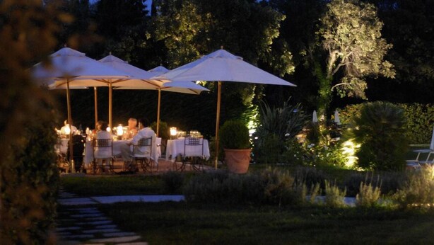 Un área de comedor al aire libre con mesas y sombrillas, rodeada de follaje y árboles.