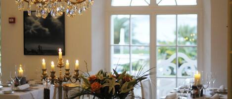 A dining table set with candles, a chandelier, and a window with a view of greenery.