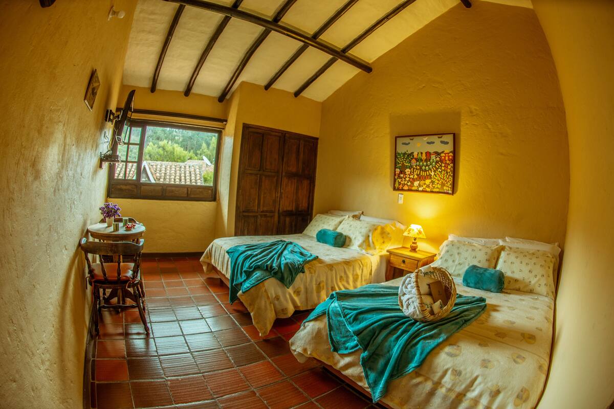 Una habitación con dos camas, una mesita con una silla, un ventanal y un techo con vigas de madera.