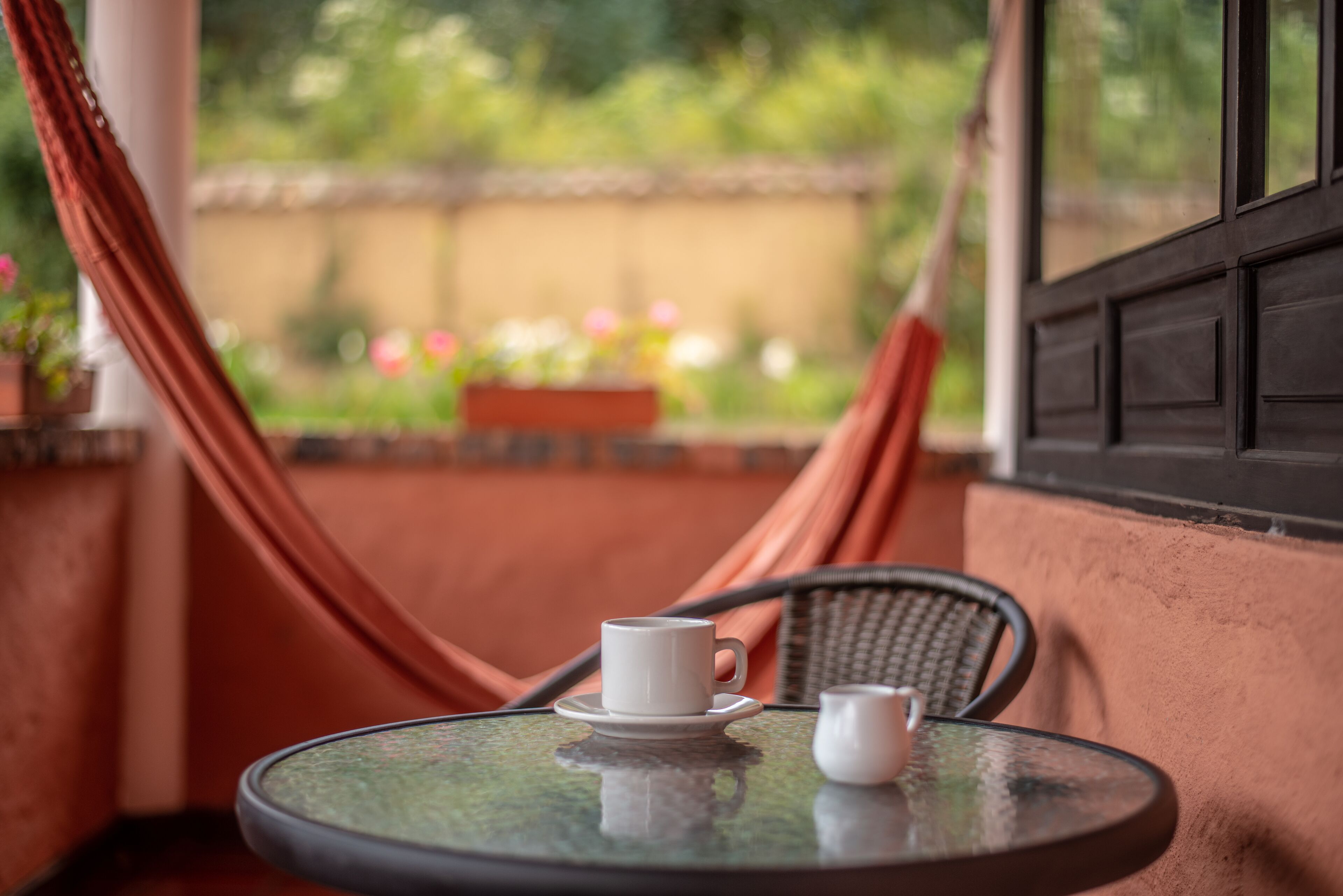 A round table with two white cups on a glass top, a hammock, and a view of a garden with flowers.