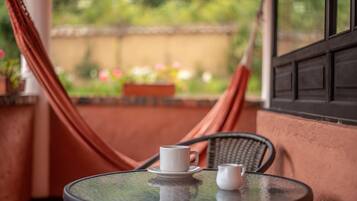 A round table with two white cups on a glass top, a hammock, and a view of a garden with flowers.