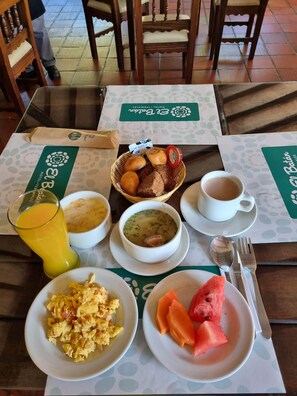Una mesa de desayuno con diversos platos, como huevos revueltos, tostadas, sopa y fruta, además de un vaso de zumo de naranja y una taza de café.