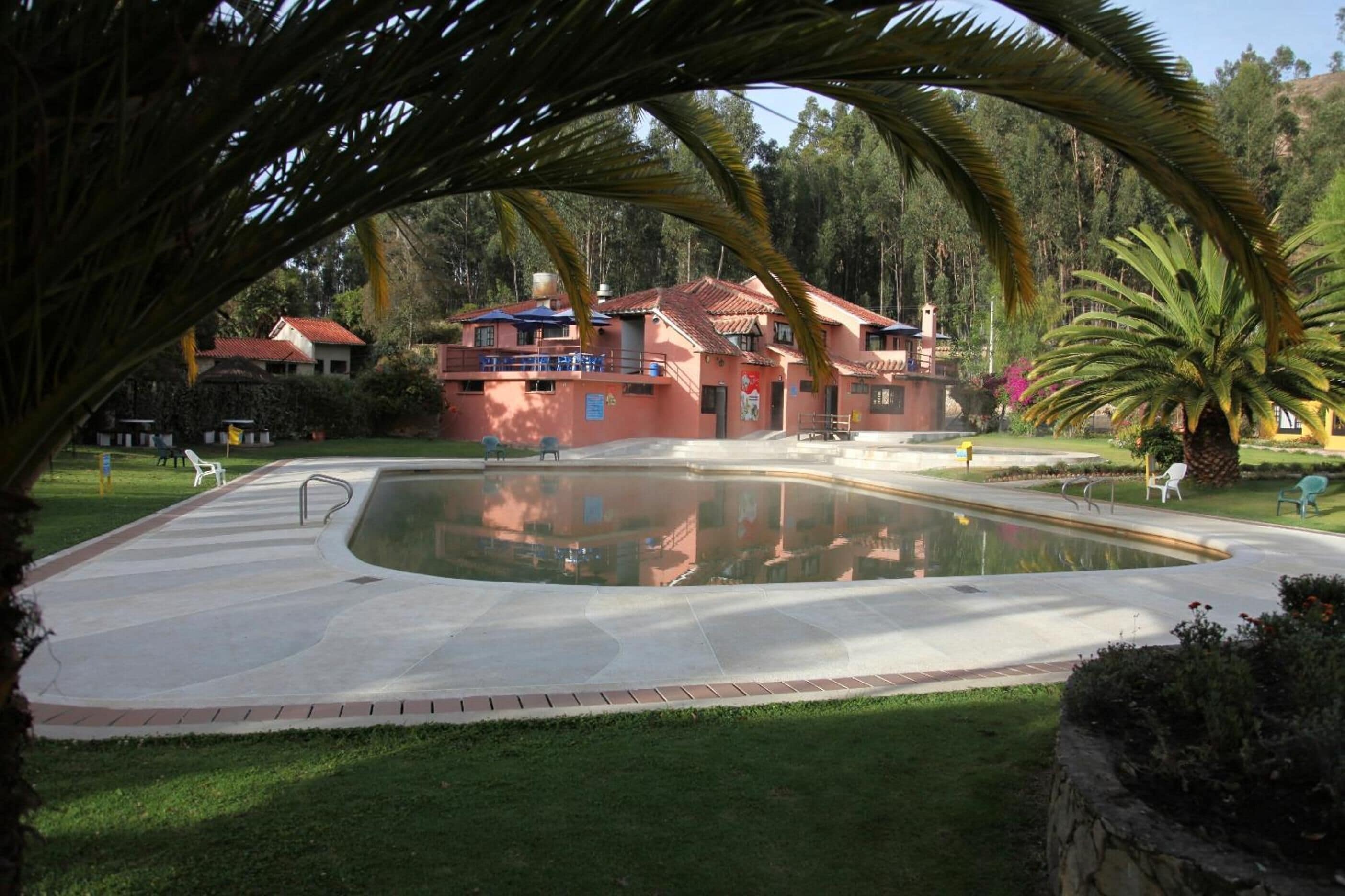 Una gran piscina con un deck circundante, un edificio rojo con techo de tejas y vegetación exuberante al fondo.