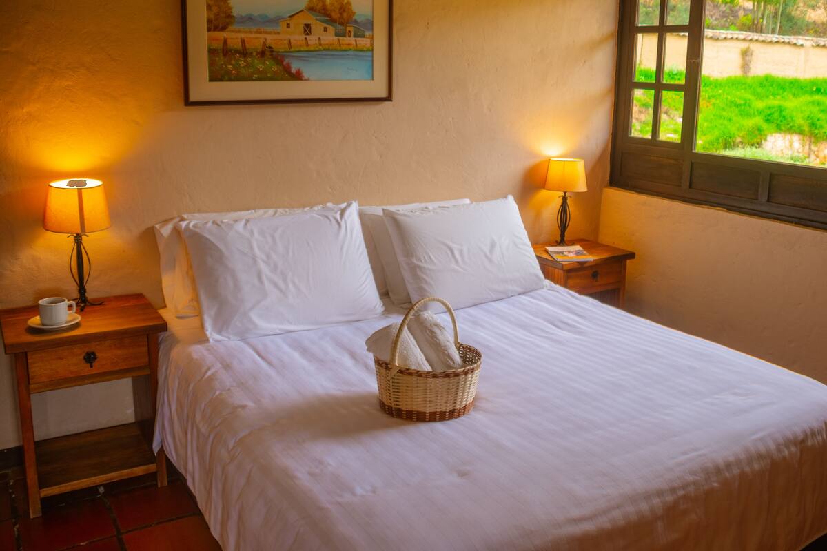 Una cama bien hecha con ropa blanca, dos lámparas de noche, una mesita de noche con una taza y un platillo, y una cesta con toallas.