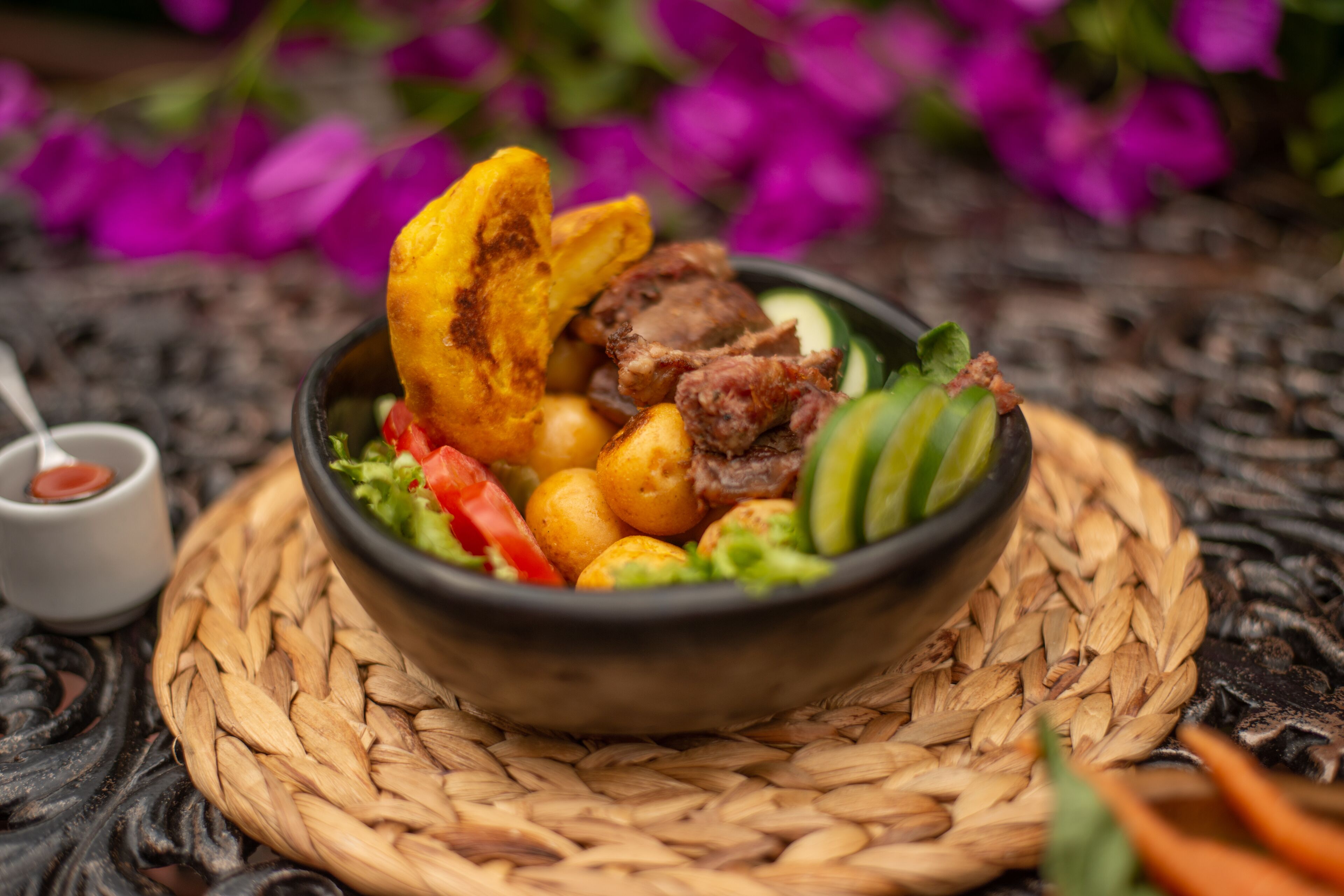 A bowl of food with various items including meat, potatoes, and vegetables, placed on a woven mat.