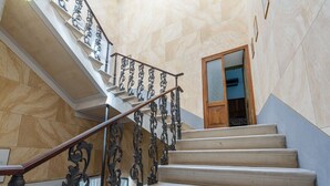 Uma escadaria com grades pretas ornamentadas e degraus bege, conduzindo a um quarto com porta de madeira e janela estampada.