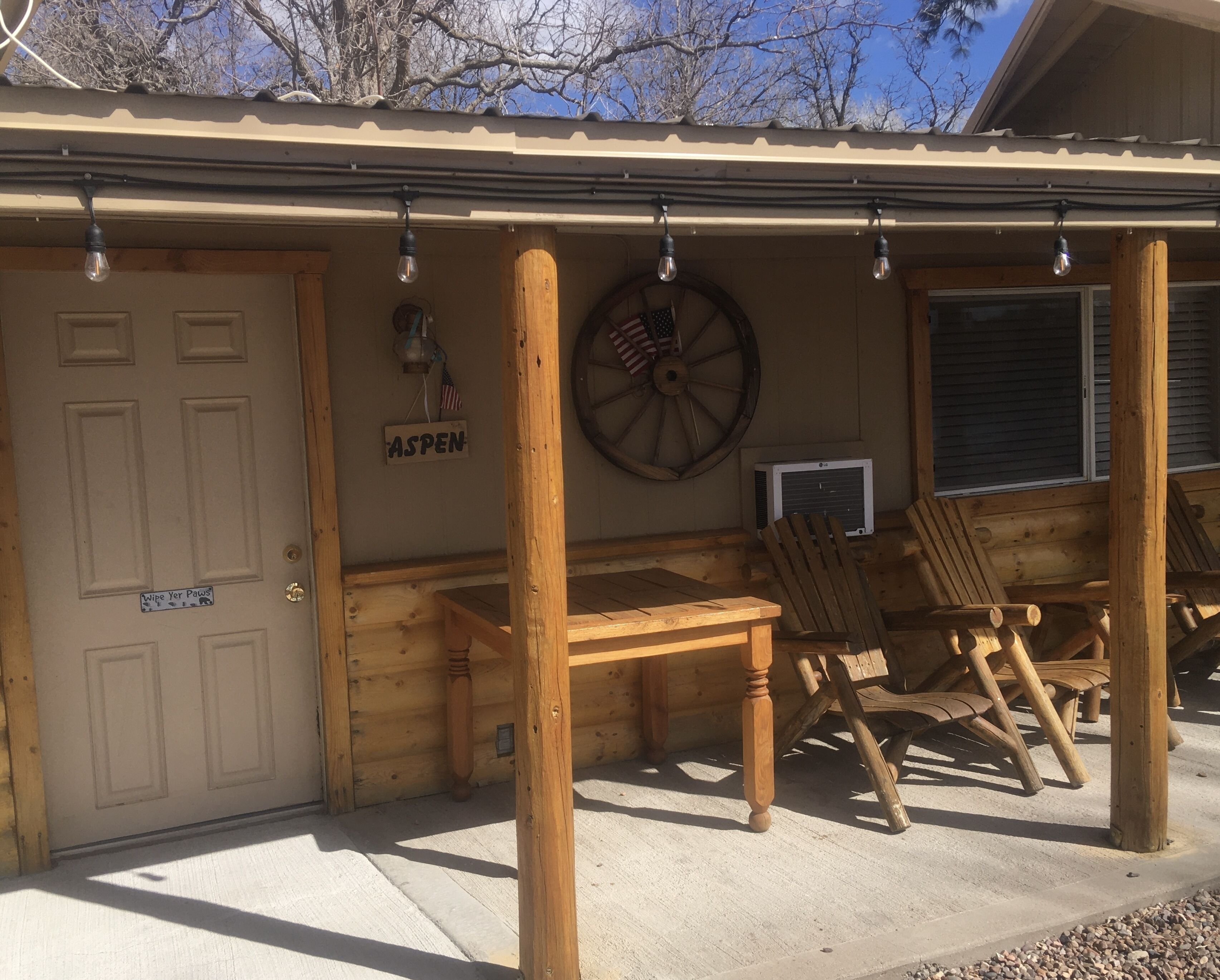 A wooden cabin with a door labeled "ASPEN," a wooden table, two wooden chairs, a mounted American flag, and a large wheel on the wall.