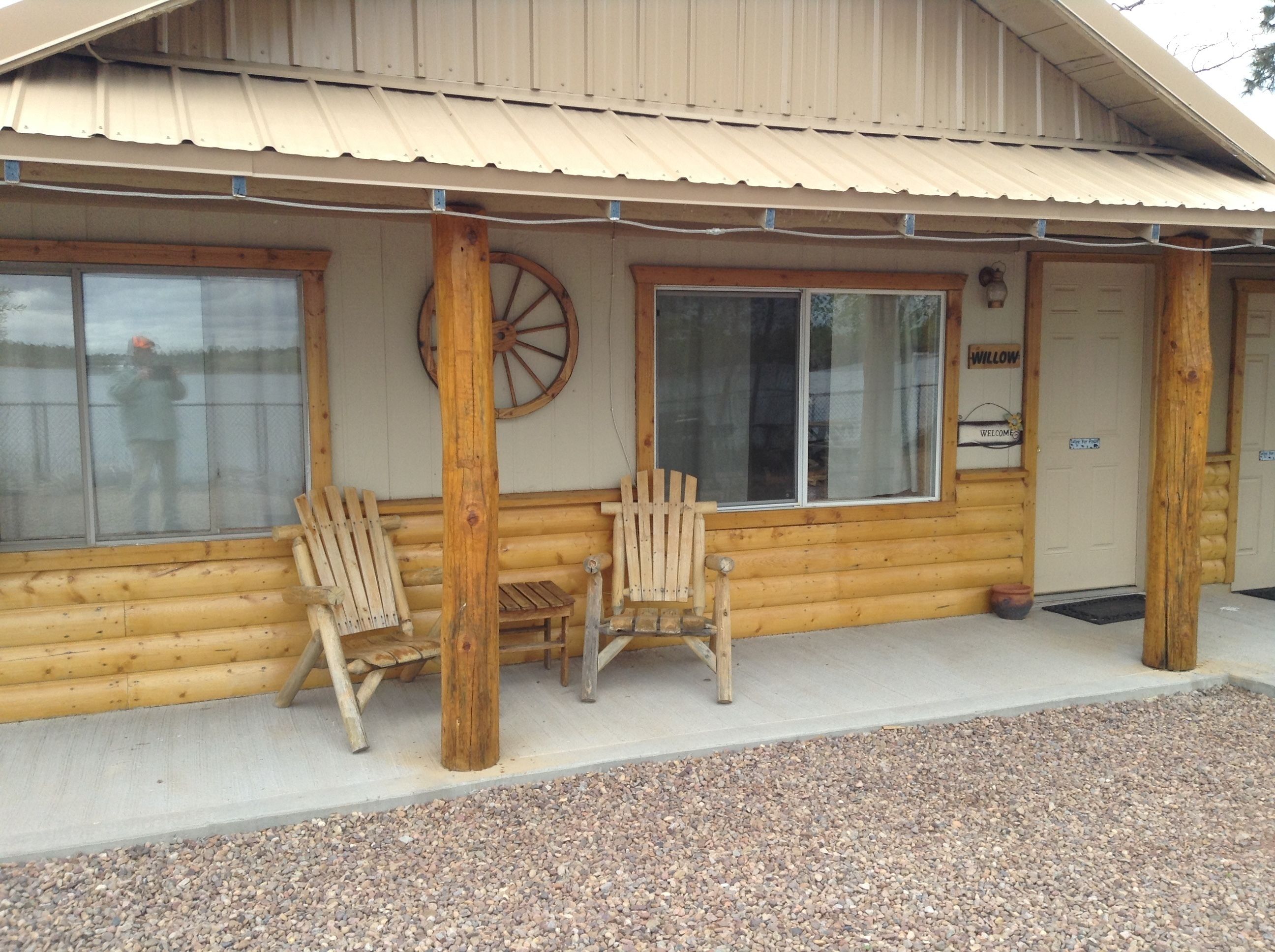 A log cabin with a porch, two Adirondack chairs, a wooden wheel, and a welcome sign.