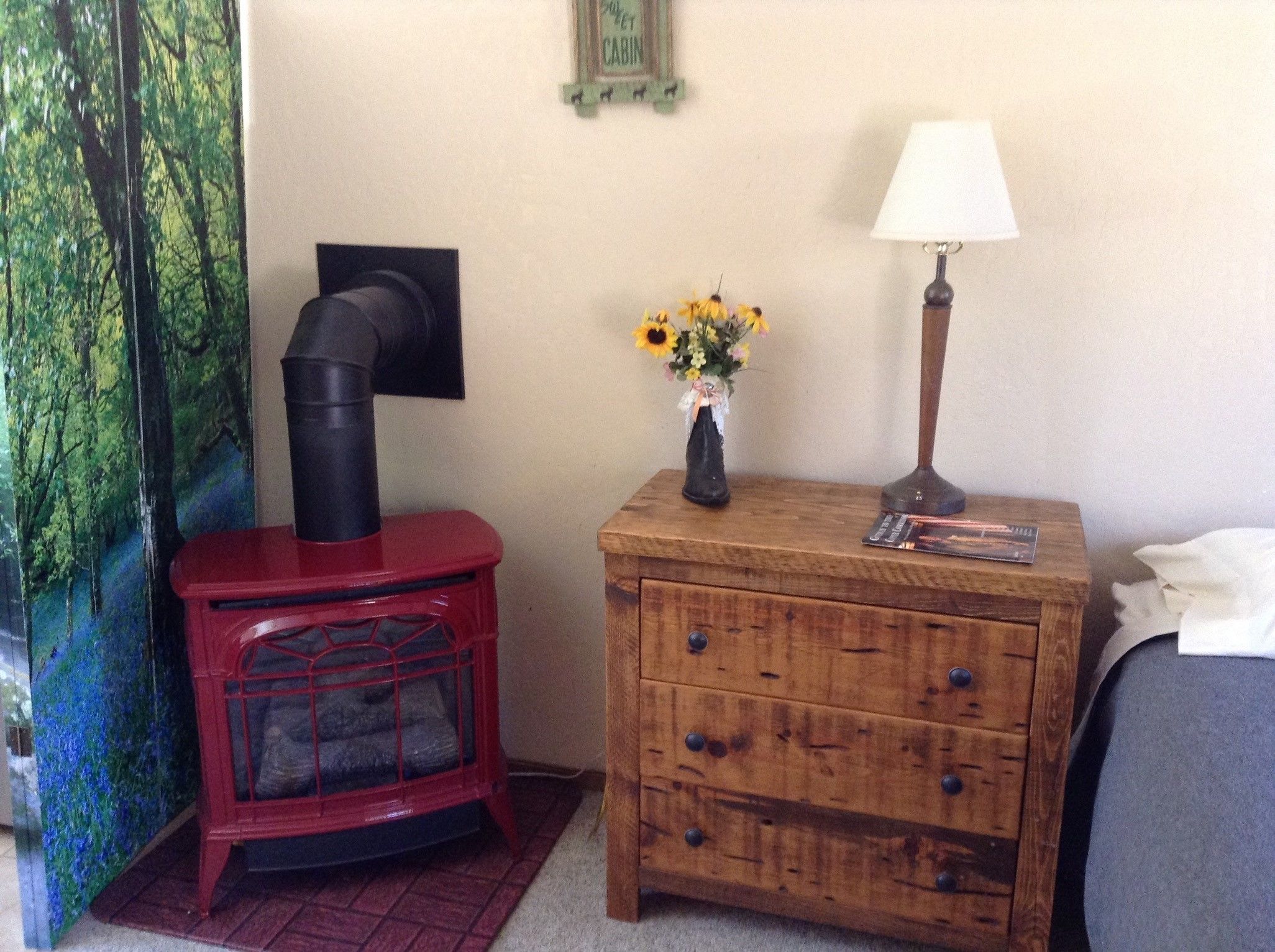 Ein Zimmer mit einem roten Kaminofen, einem Nachttisch aus Holz mit einer Lampe und einer Blumenvase sowie einem Bett mit weißer Bettdecke.