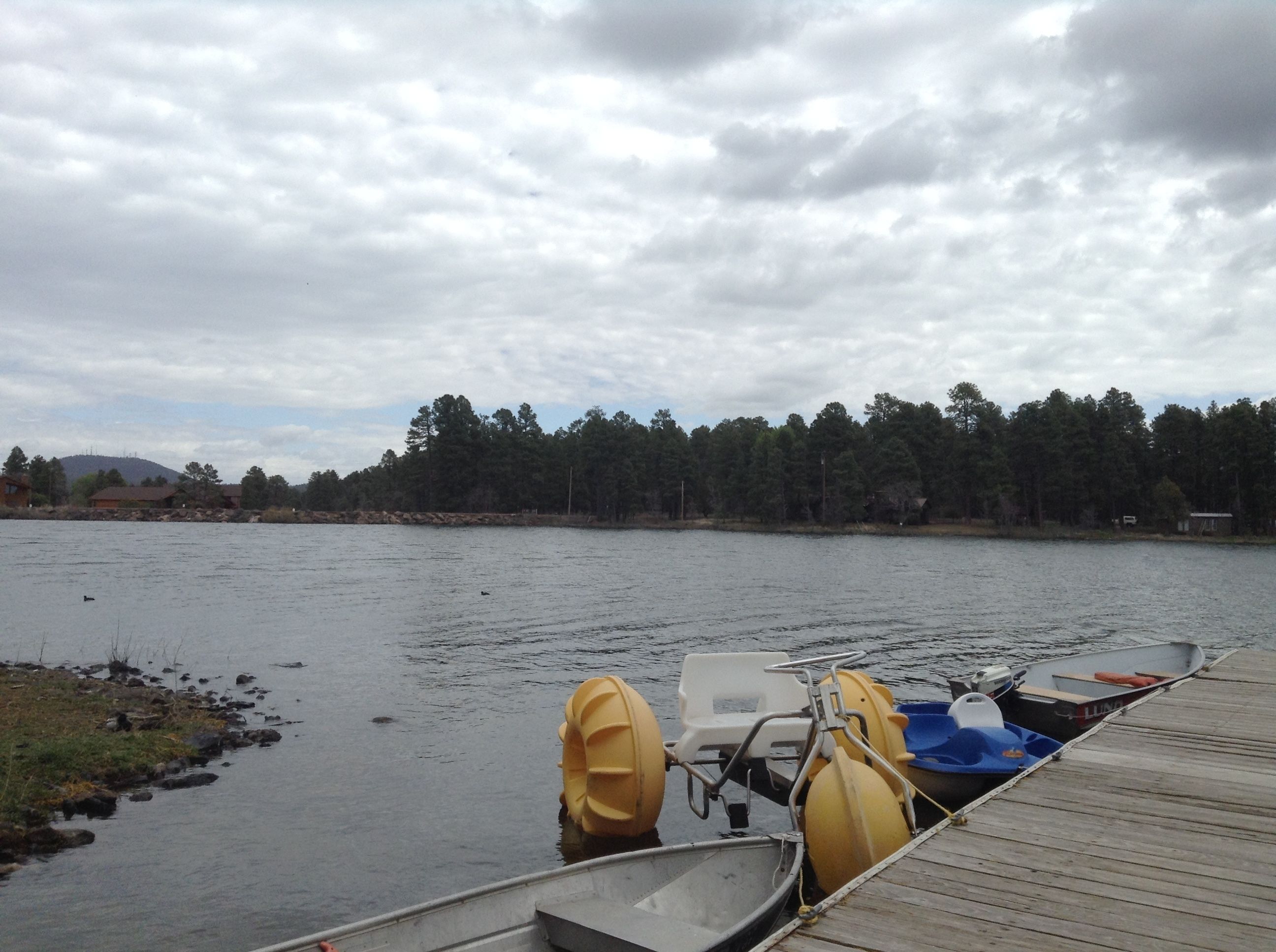 A calm lake with a wooden dock, a rowboat, and a motorboat with yellow floats.