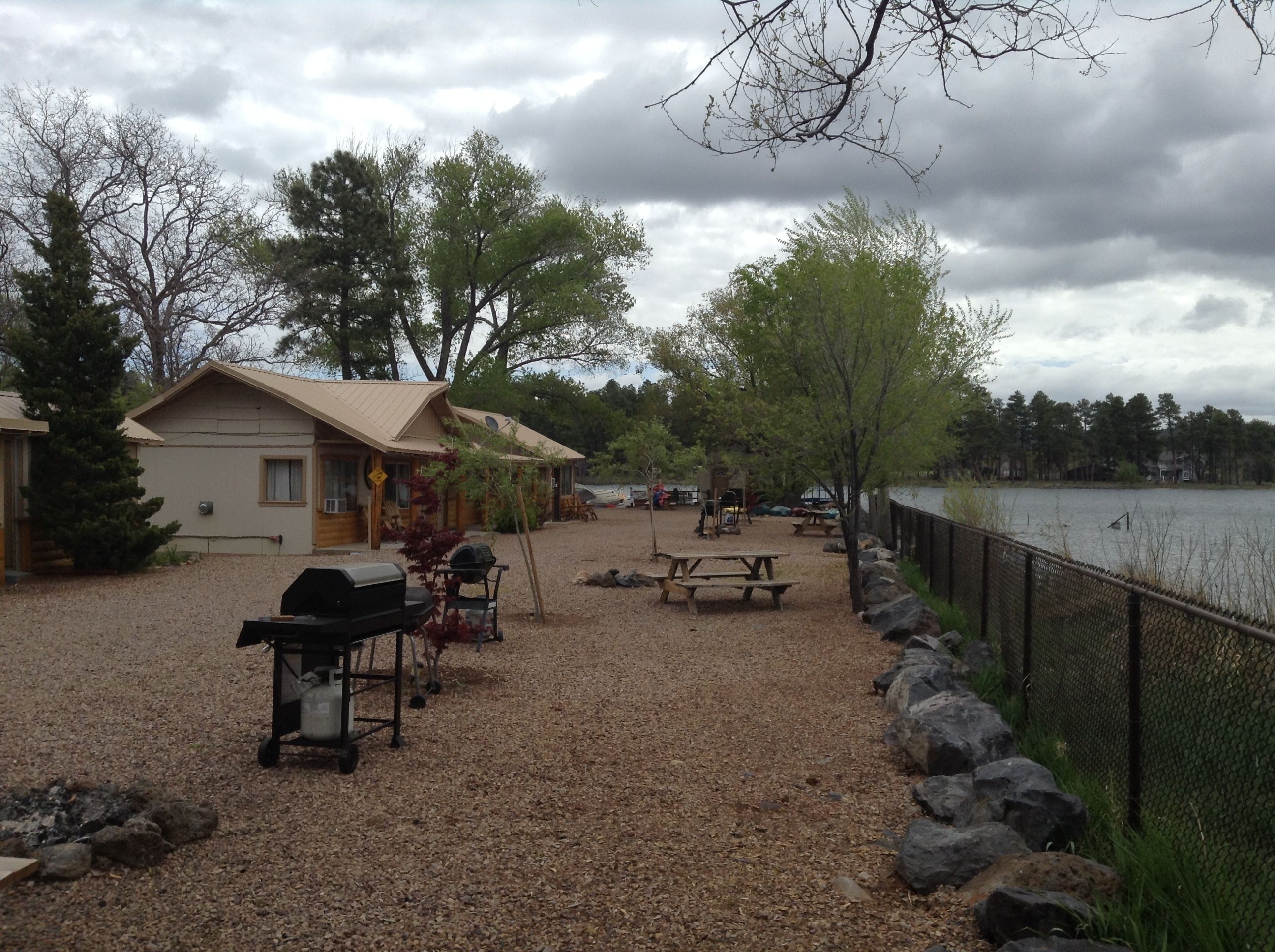 Una cabaña junto al lago con un patio de grava, mesas de picnic y una parrilla.