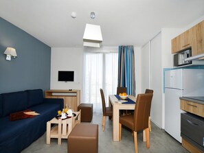 A modern living room with a sofa, a dining table with chairs, a kitchen area with appliances, and a television on the wall.