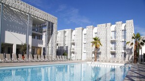 A swimming pool with a clear blue water surface, surrounded by white buildings with geometric patterns, and a clear sky above. - Apartment in France with Furnished Terrace (Cap d'Agde)