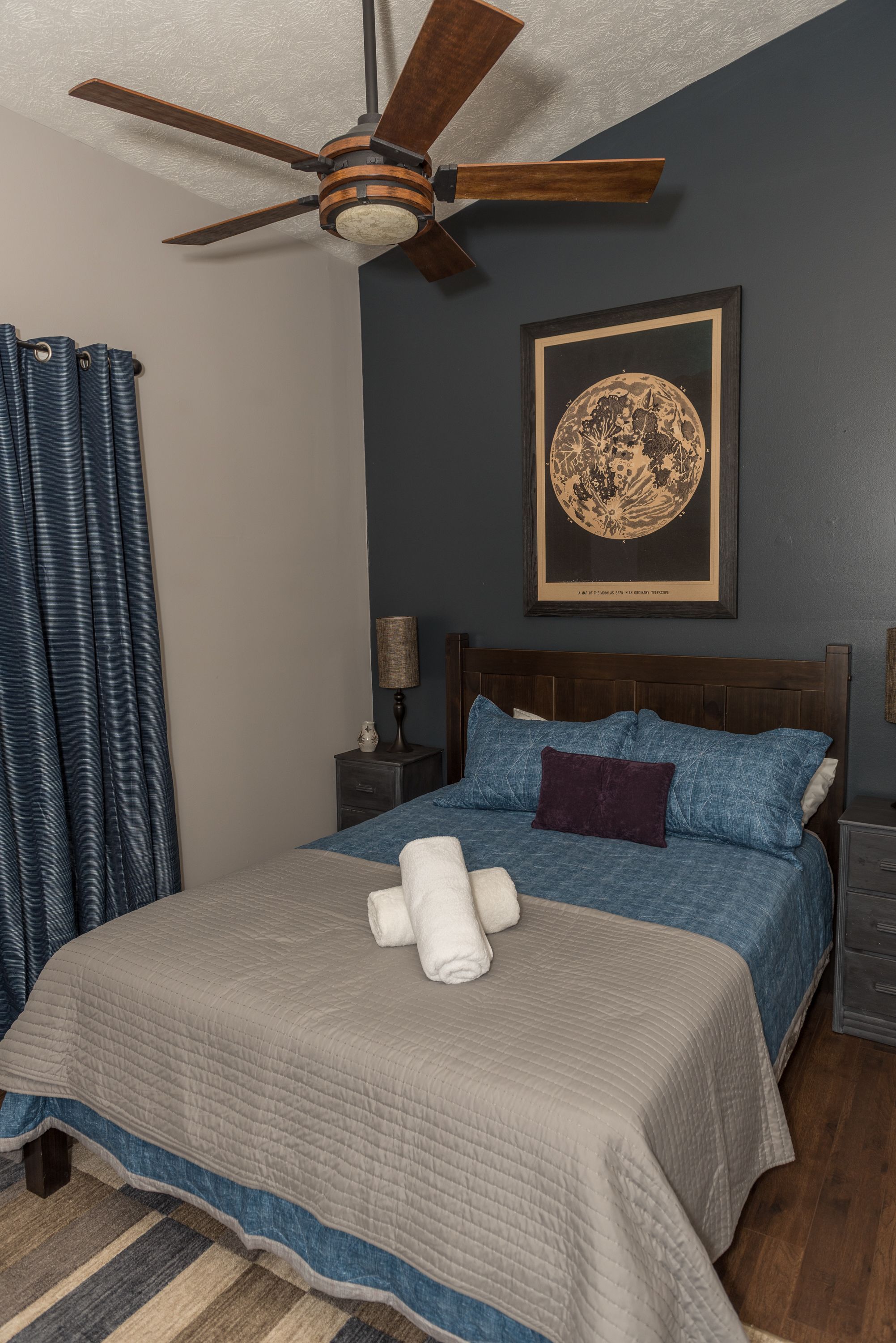 A bedroom with a bed, a ceiling fan, a nightstand, a lamp, and a framed artwork on the wall.