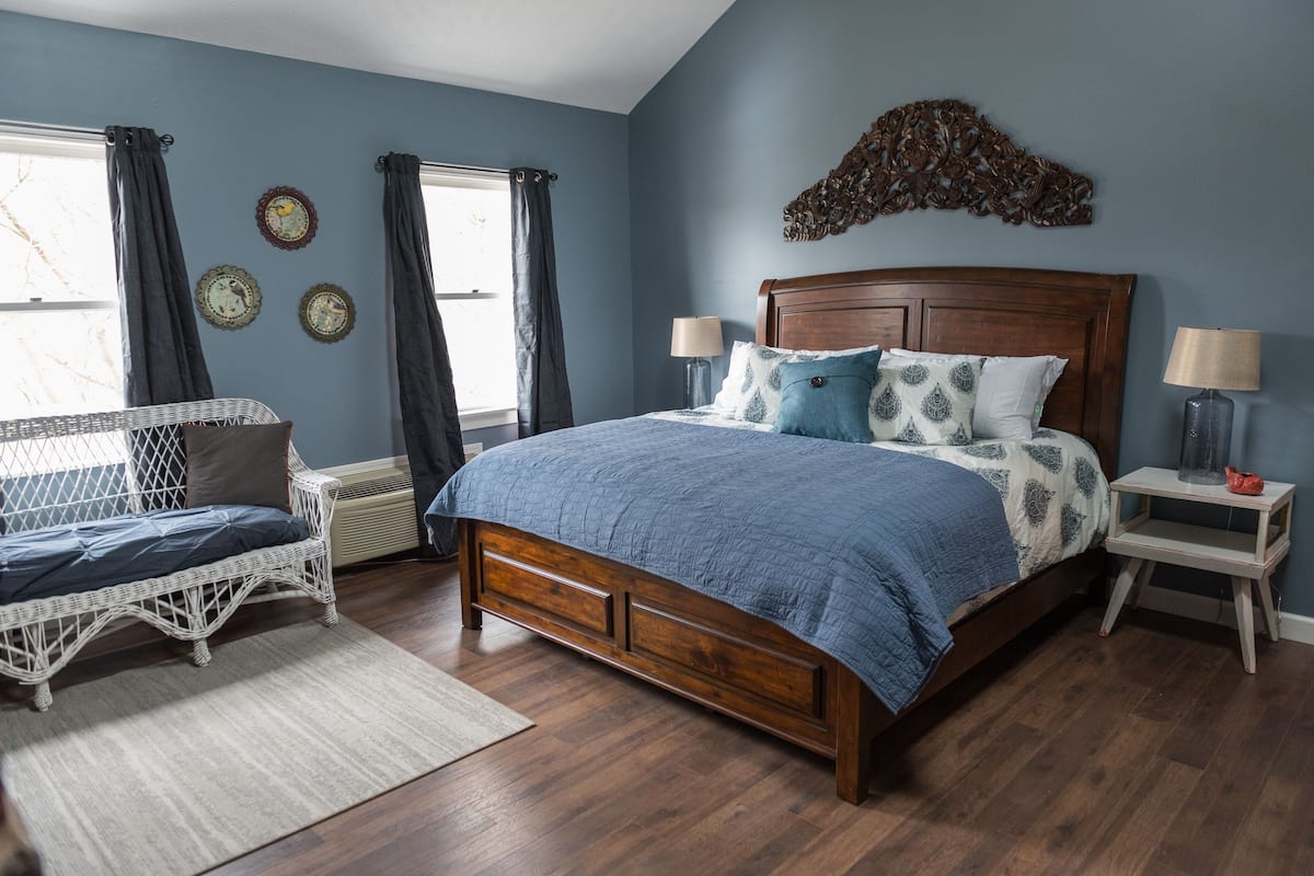 A bedroom with a wooden bed, two bedside tables, a wicker chair, and a window with curtains.