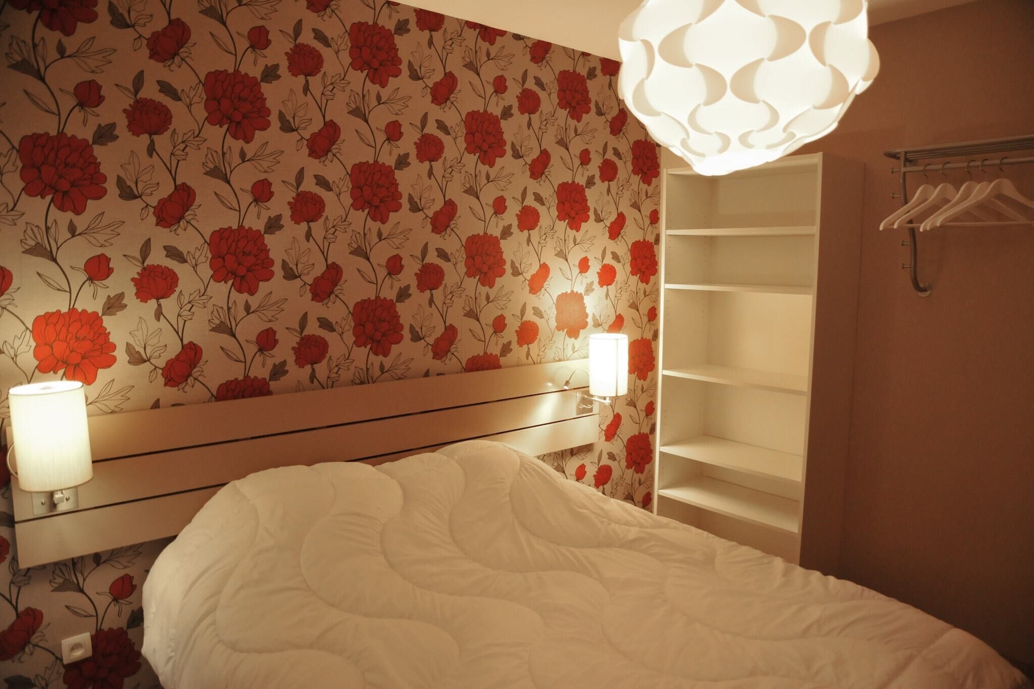 Une chambre d’hôtel avec un papier peint à motifs floraux, un lit avec une literie blanche, une lampe de chevet et une armoire avec une tringle pour suspendre les vêtements.