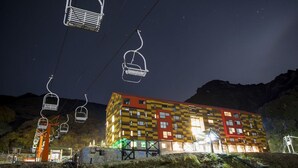 Un edificio de varios pisos con fachada a cuadros, ventanas iluminadas y un telesilla al fondo.
