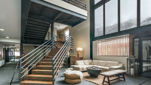 Un salón amplio con una escalera, un sofá, una mesa de centro y un gran ventanal con vistas a un paisaje nevado.