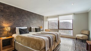 Un dormitorio con una cama grande, cabecera de madera, mesita de noche, una silla y un ventanal con vistas a un área boscosa.