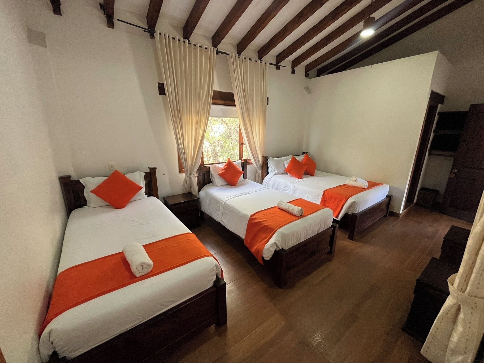 Et hotelværelse med tre enkeltsenge, alle med hvide sengetæpper og orange detaljer.