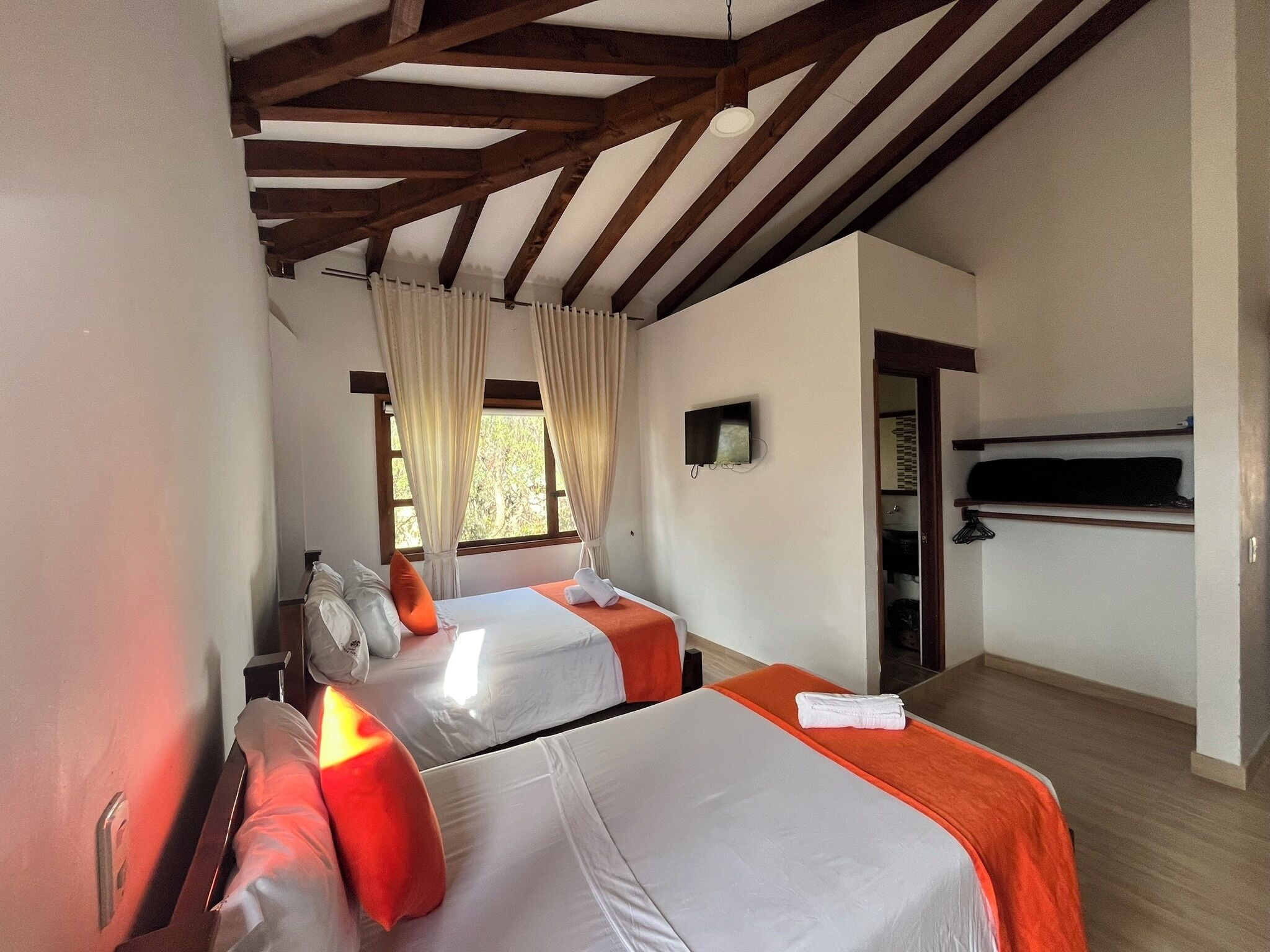 A hotel room with two beds, wooden beams on the ceiling, a TV on the wall, and a window with curtains.