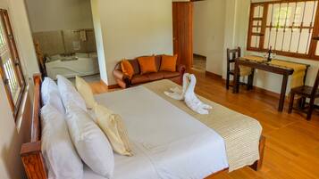 Habitación de hotel con una cama grande, piso de madera, un sofá, una mesa con una botella y una ventana con barrotes.