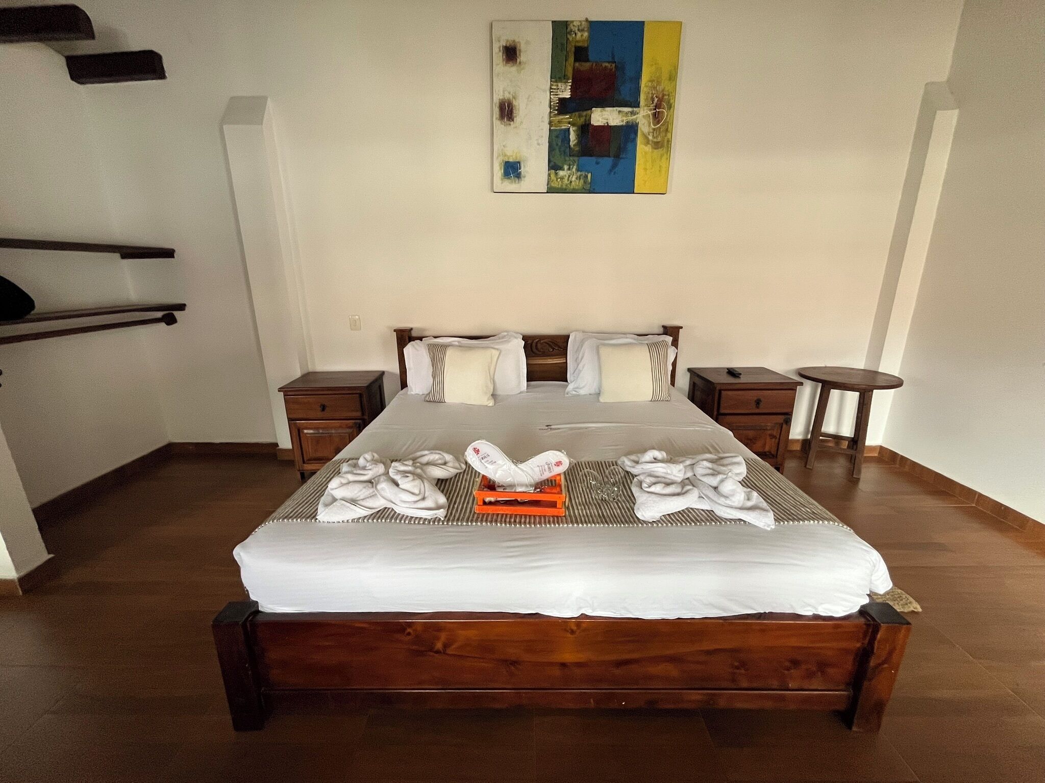 A neatly made bed with white linens, two bedside tables, a small round table, and a painting on the wall.