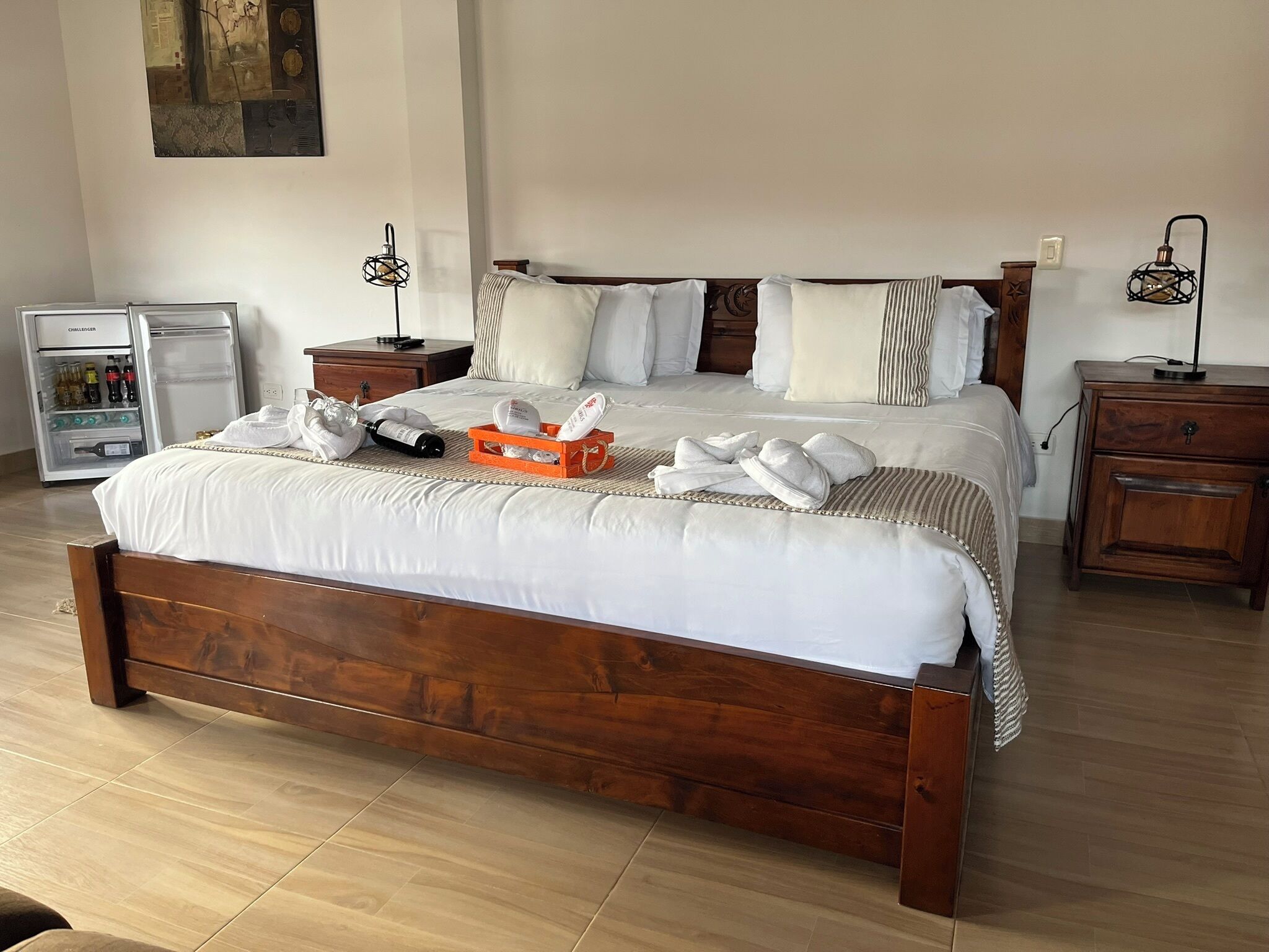 Una cama bien tendida con sábanas blancas, cabecera de madera y una pequeña bandeja naranja con artículos de aseo. Hay un minibar a la izquierda y una mesita de noche con lámpara a la derecha.