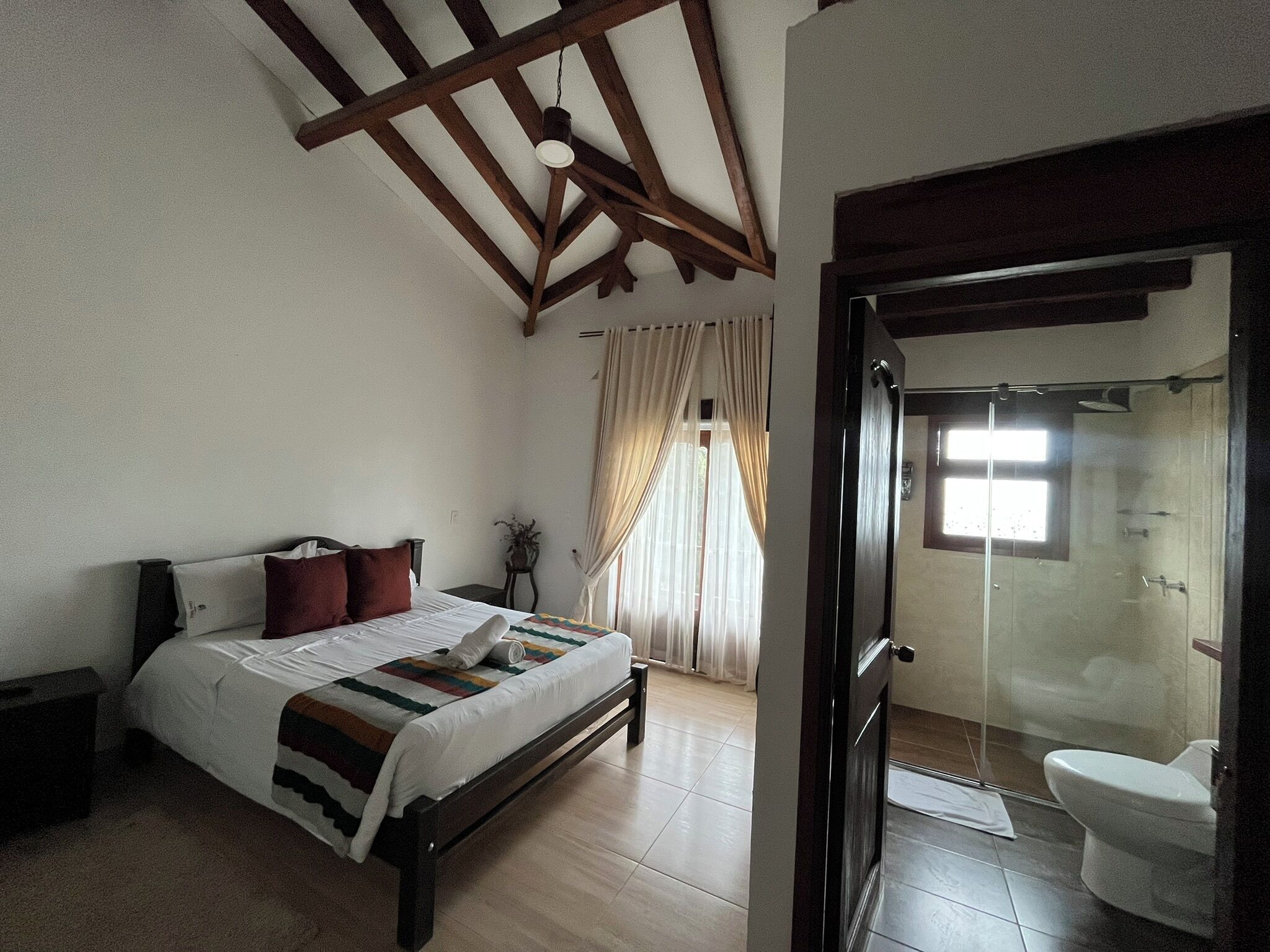 A bedroom with a bed, a bedside table, a window with curtains, and a view of a bathroom with a shower and toilet.