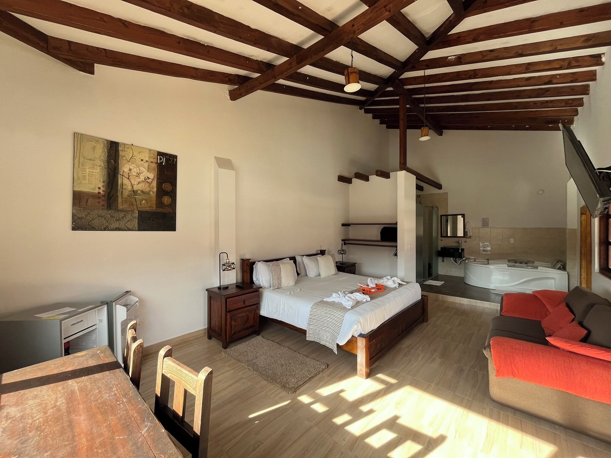 A bedroom with a bed, bedside table, and a desk. There is a chair, a sofa, and a bathroom with a bathtub.
