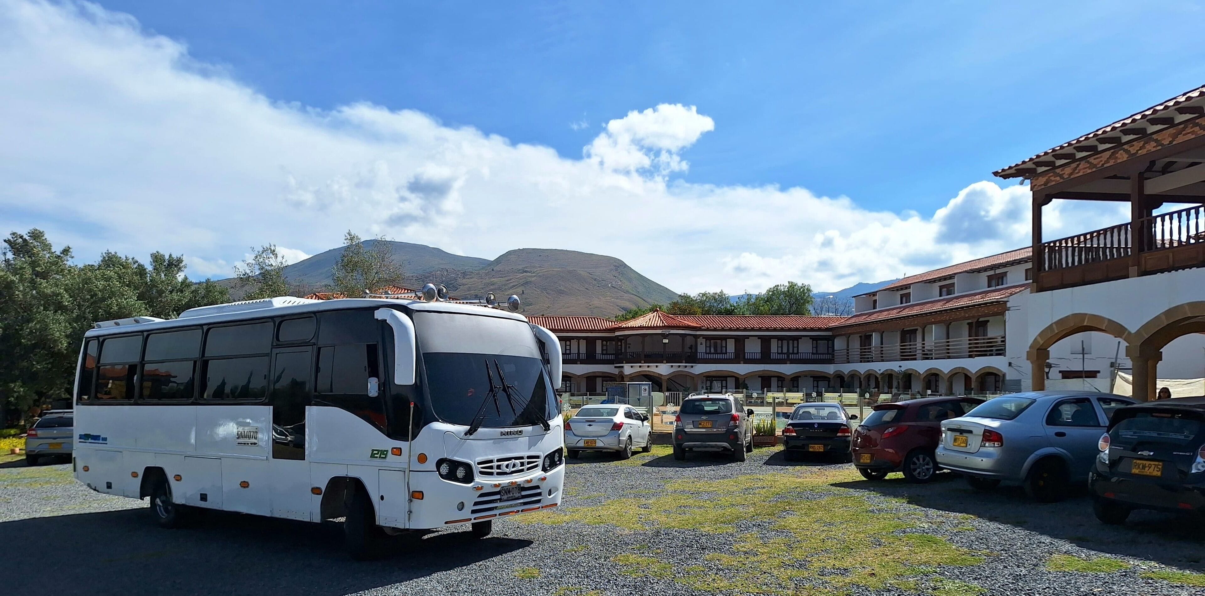 Un autobus est garé devant un immeuble, avec plusieurs voitures à proximité, un ciel dégagé et des montagnes en arrière-plan.