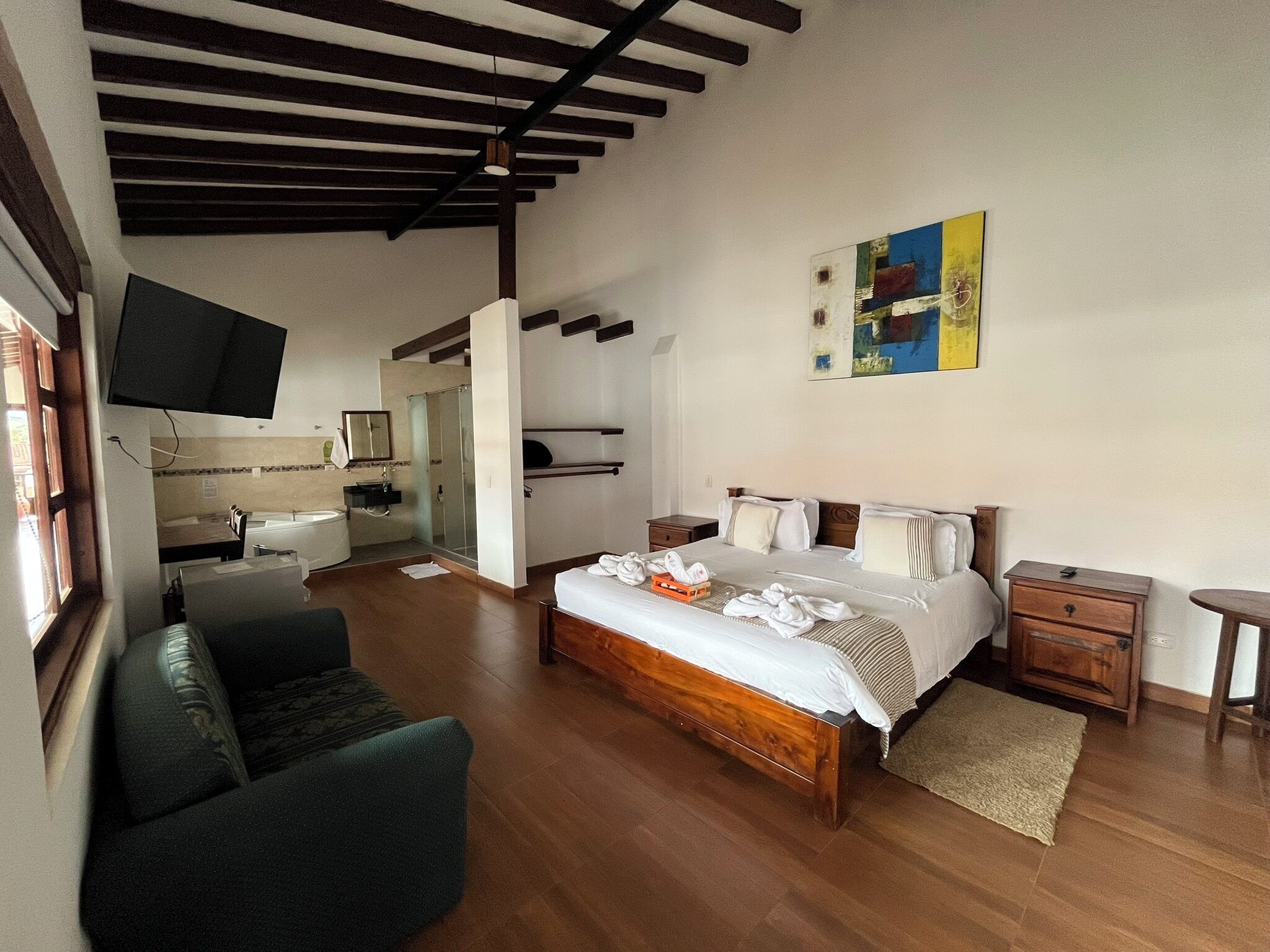 Una habitación de hotel con una cama de madera, un sillón verde, un televisor, un baño con ducha y un cuadro en la pared.
