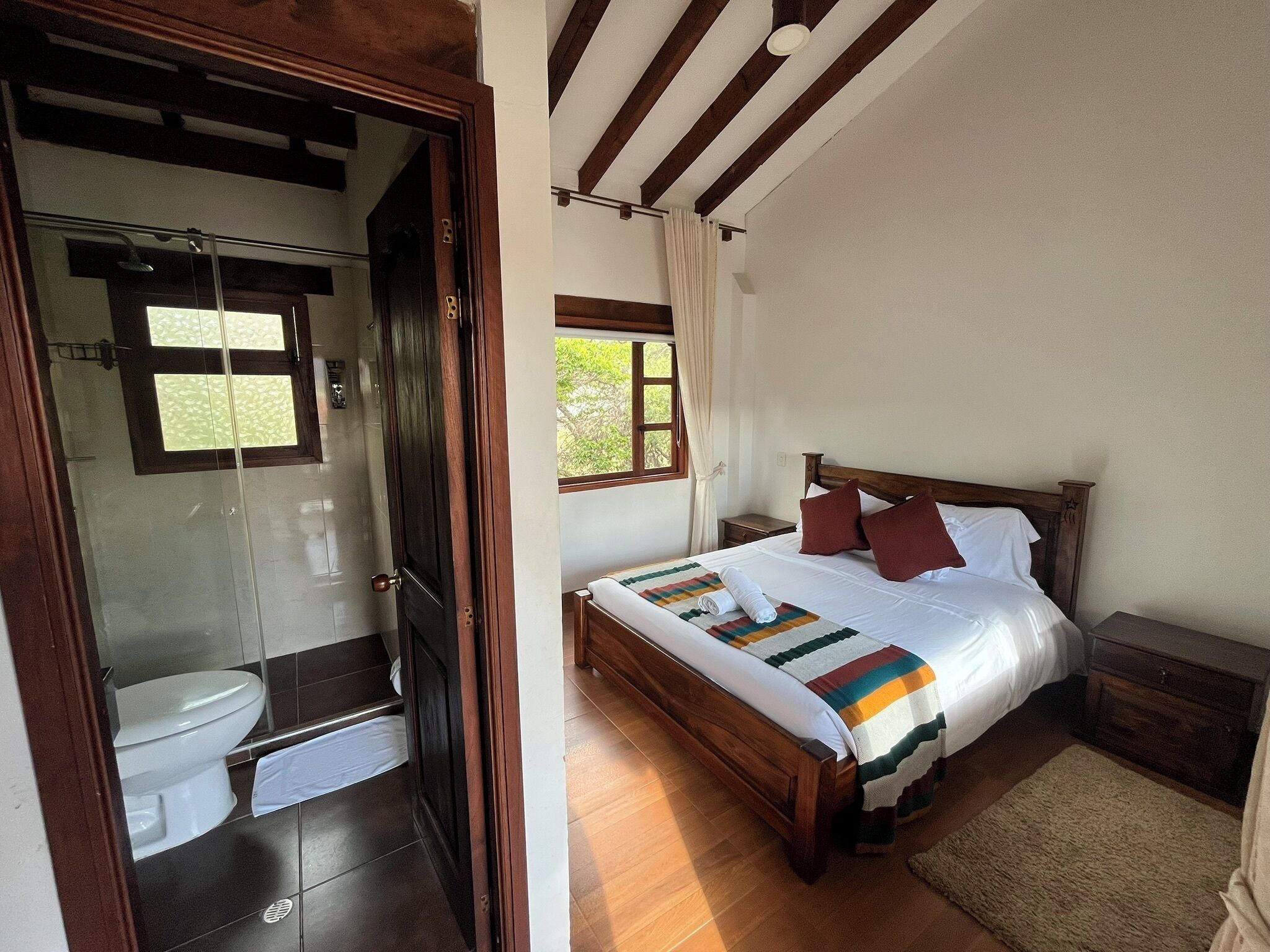 Une chambre à coucher avec un lit en bois, une table de chevet, une fenêtre avec des rideaux et une salle de bain équipée de toilettes et d’une douche.