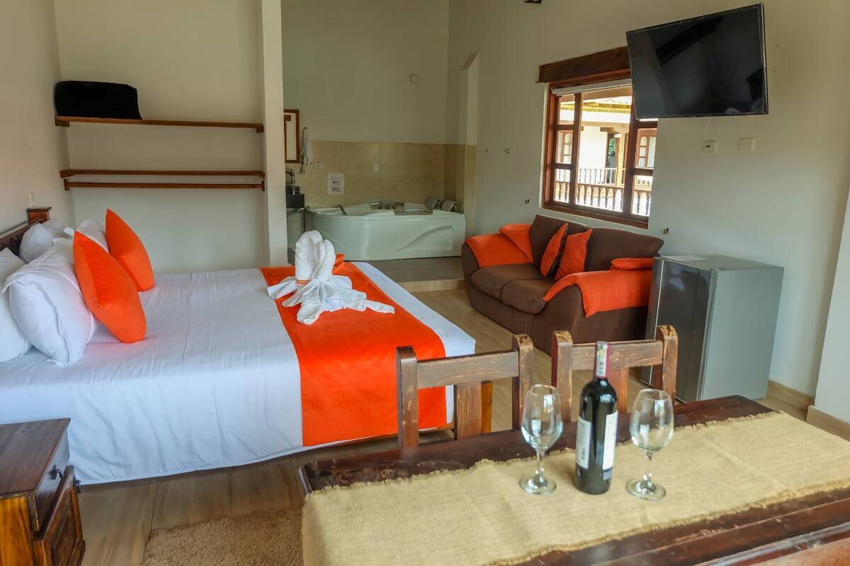 A hotel room with a bed, a sofa, a TV, and a table with wine glasses and a bottle.