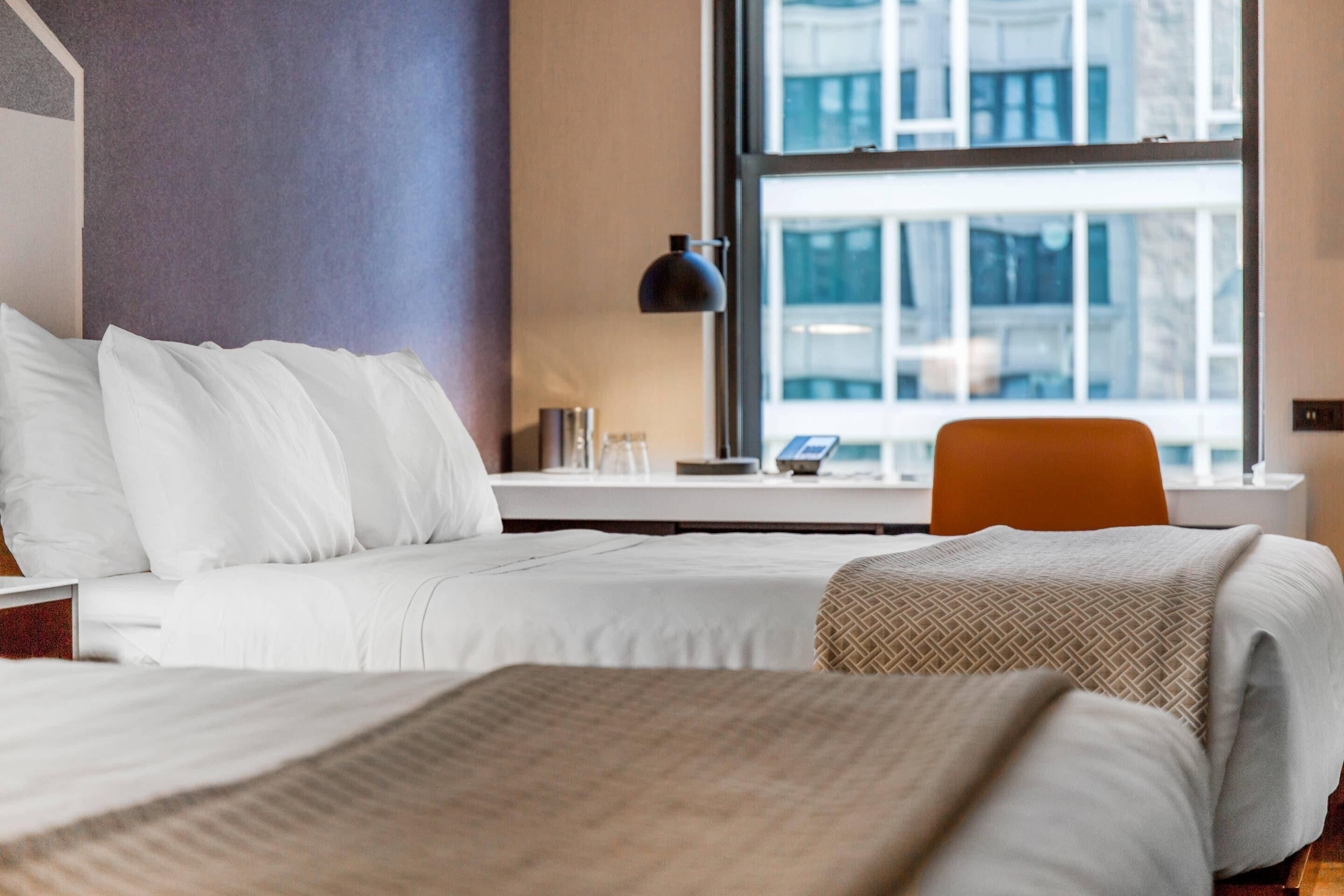 A hotel room with two beds, a desk, a chair, and a large window with a view of buildings.