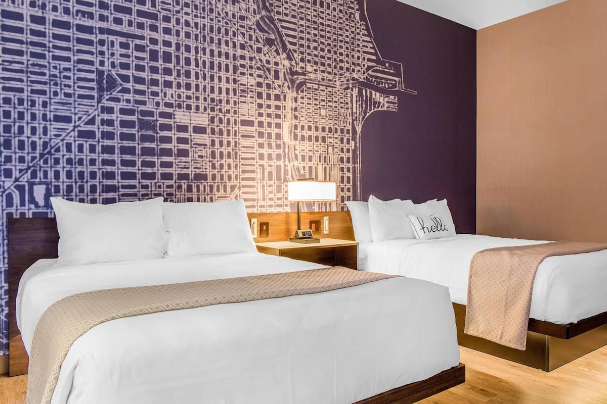 Une chambre d’hôtel avec deux lits, une table de chevet avec une lampe et un mur orné d’une fresque représentant un paysage urbain.