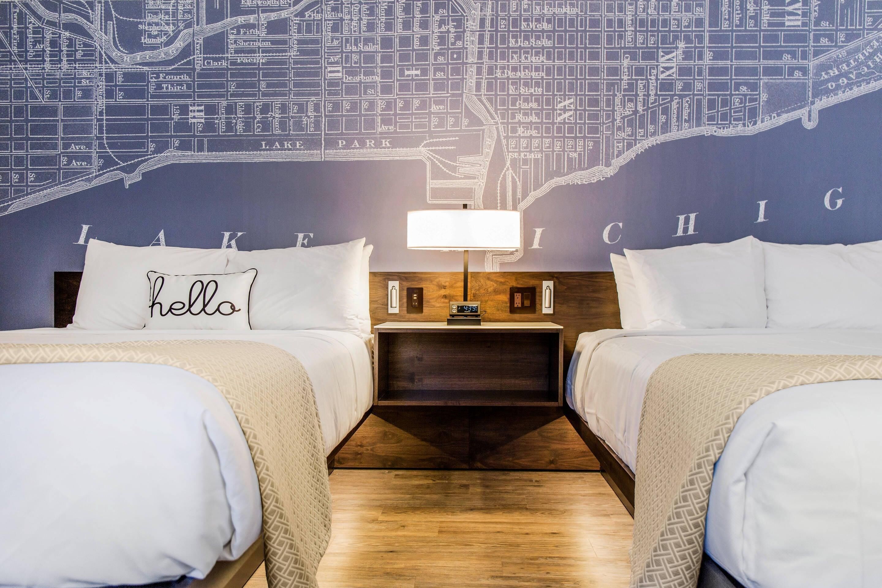 Una camera d'albergo con due letti, un comodino con una lampada, una mappa appesa al muro e un cuscino con la scritta 'hello'.